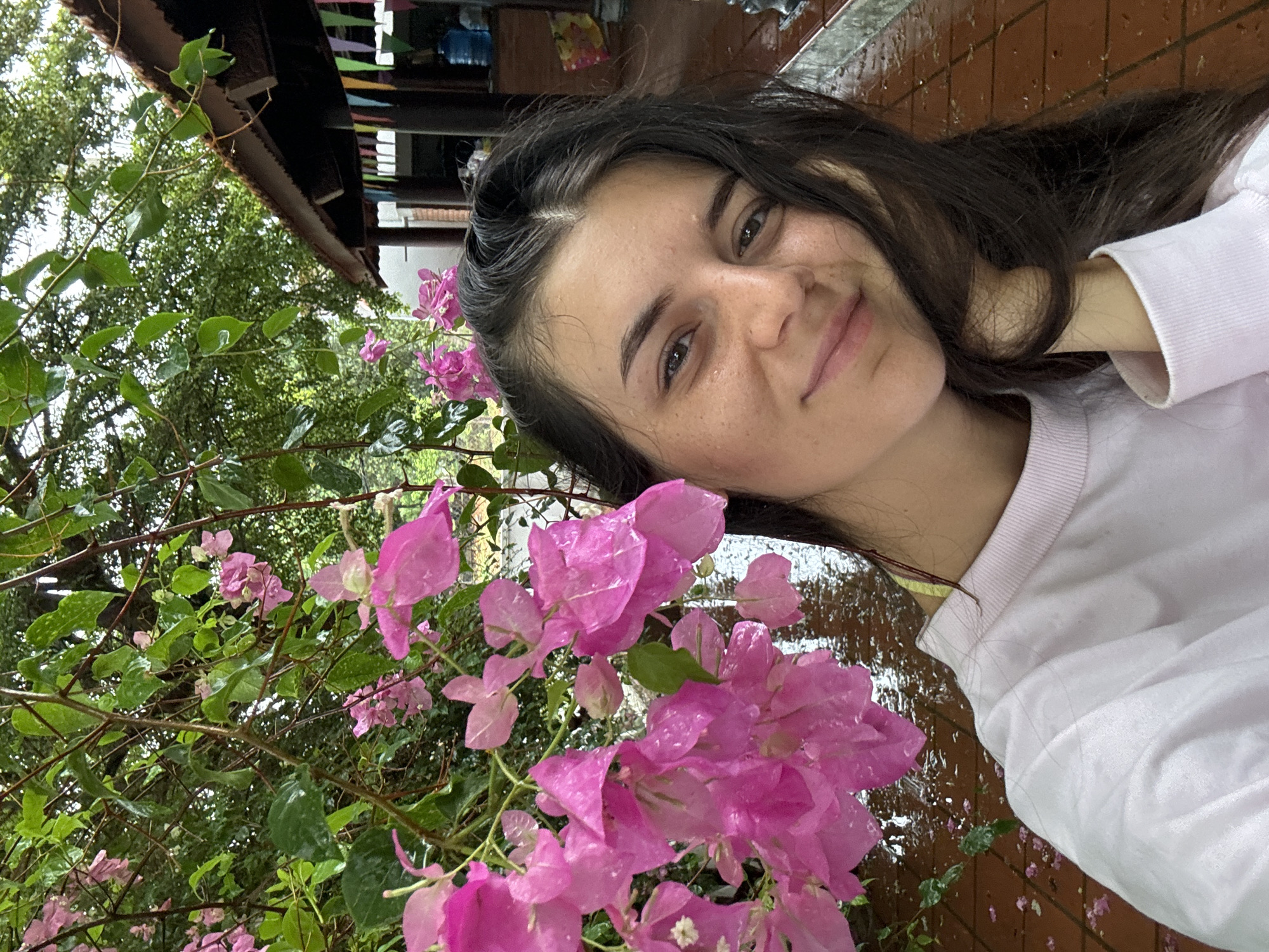 A young woman with long, dark hair is posing for a selfie next to a blooming bougainvillea bush. The flowers are a vibrant pink and appear to be wet, suggesting recent rain. The woman is wearing a light-colored sweatshirt and has her head tilted slightly, smiling at the camera.
The background includes greenery, possibly trees, and a building with a roof visible at the top of the frame. The ground appears to be tiled and wet, with scattered fallen petals.
Given the presence of bougainvillea and the architectural style of the building visible in the background, the location is likely somewhere in a tropical or subtropical region. While specific details are limited, based on the prompt, the location is Mũi Né, Vietnam.