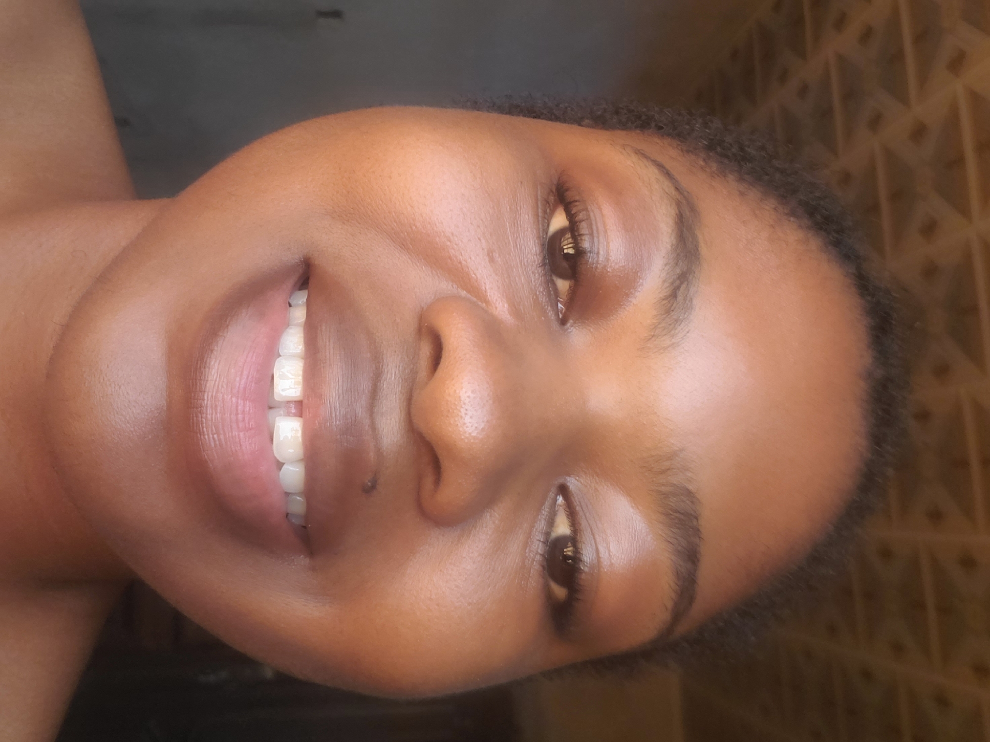 A close-up, front-facing portrait of a young adult female with a rich brown skin tone, captured in Kakau, Nigeria. The subject is smiling broadly, revealing her upper teeth, which show a noticeable diastema between the two central incisors. Her dark, natural curly hair frames her face. She has dark brown eyes with visible dark eyelashes and well-defined, natural eyebrows. A small dark mole is present below her right nostril. Her lips are full and have a natural sheen. The upper background features a textured surface with a repeating geometric pattern, consistent with a decorative ceiling or wall panel, in a warm brown hue. The lower background is dark and indistinct, suggesting an indoor setting with uneven lighting. The subject's bare shoulders are visible in the lower frame.