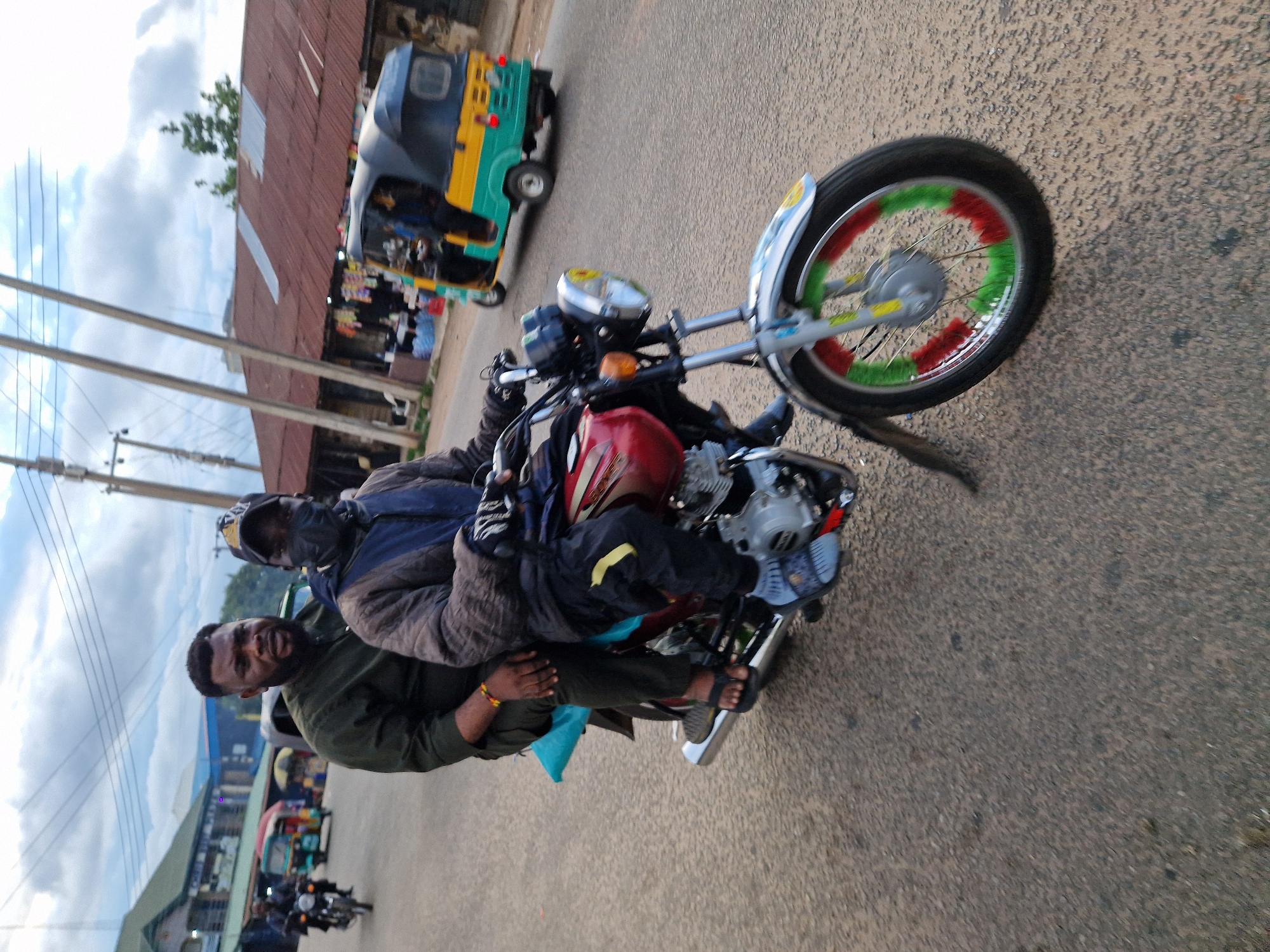 In Jos, Nigeria, on a cloudy day, two men are riding a red motorcycle on a paved road. The man in the front, wearing a dark green long-sleeved shirt and dark pants, has a beard and is looking forward. The man behind him, wearing a dark hooded jacket, a blue under-layer, dark pants with a yellow stripe, and a black face mask, is holding onto the driver. The motorcycle has a silver exhaust pipe and a front wheel with red and green accents. In the background, a yellow and green tricycle with a blue top is moving away, and buildings with corrugated iron roofs line the street. Power lines are visible against the sky.