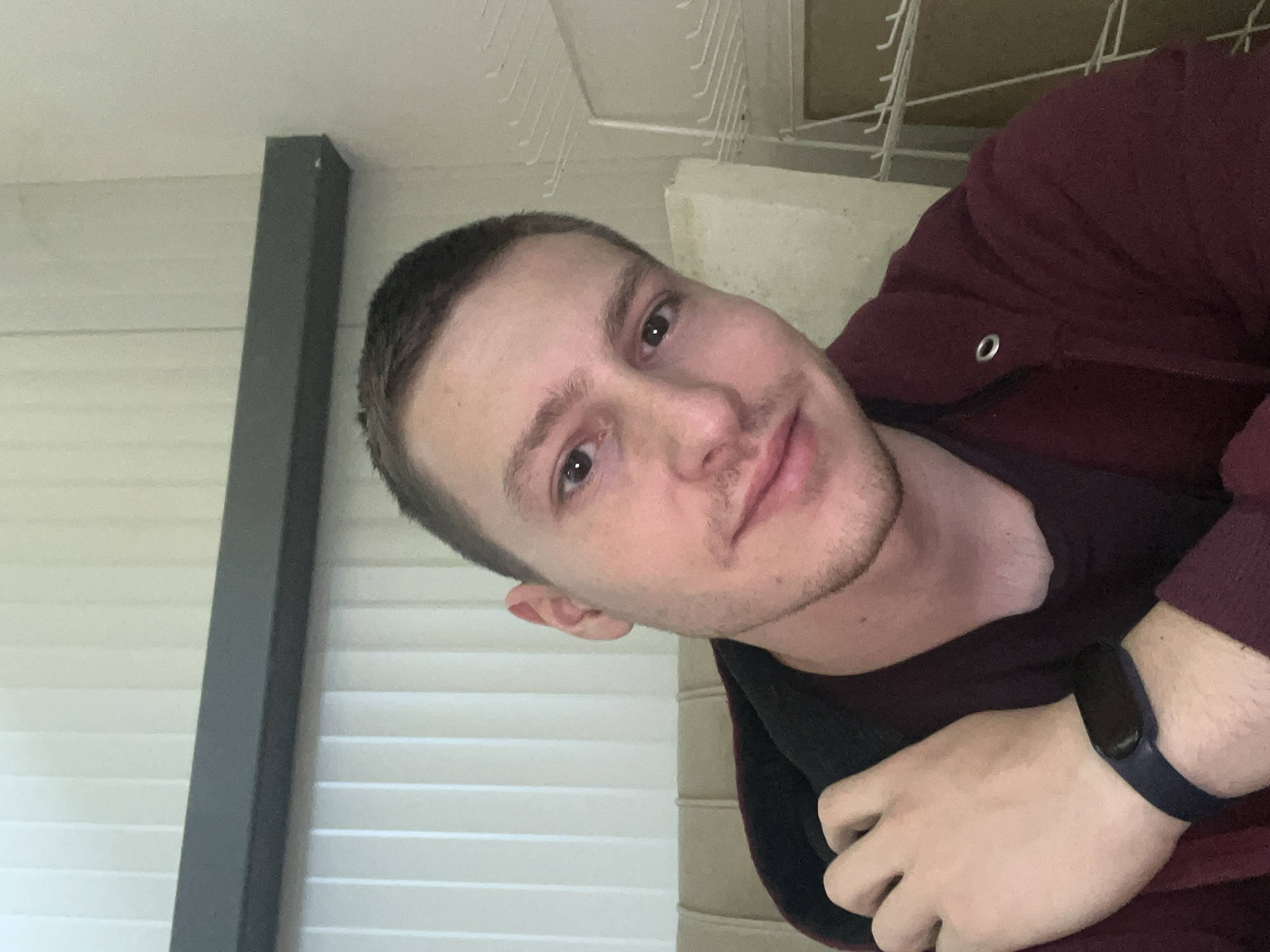 A Caucasian male with short, light-brown hair and stubble is posing in a selfie. He is wearing a maroon hooded sweatshirt and a dark-colored fitness tracker on his left wrist. The background includes white window blinds, a grey support beam, and what appears to be a white metal rack on the ceiling. There is no information in the image to determine the location is Burgas, Bulgaria.