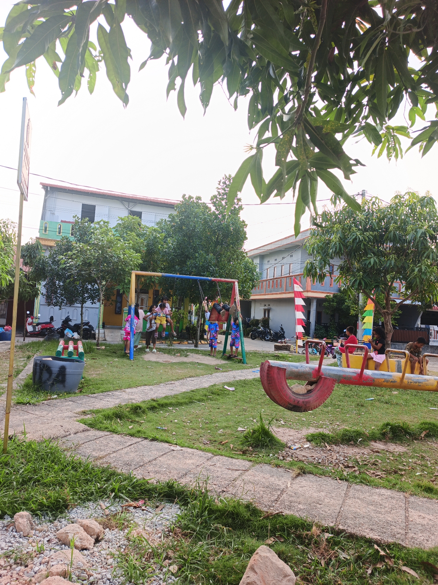 This is a medium-angle, outdoor, eye-level shot of a playground in Batam, Indonesia. The playground is situated in front of a residential area with multi-story buildings. The foreground is dominated by a path made of concrete slabs separated by grass. To the left of the path, there is a large grey dumpster with black spray paint markings, and to the right, a seesaw made from a tire and metal frame is visible.
In the midground, a swing set with colorful poles and chains is occupied by several children. Some children are swinging, while others are standing around it. Behind the swing set, trees with green foliage are scattered across the lawn. Several motorcycles are parked near the buildings in the background. The buildings themselves are painted in various colors, including light blue, yellow, and gray, with some featuring decorative elements like red and white striped flags.
The background shows the sky, which is overcast and bright, suggesting it is daytime. The overall scene depicts a community playground where children are engaging in recreational activities. The image has a casual, everyday feel, capturing a slice of local life in Batam.