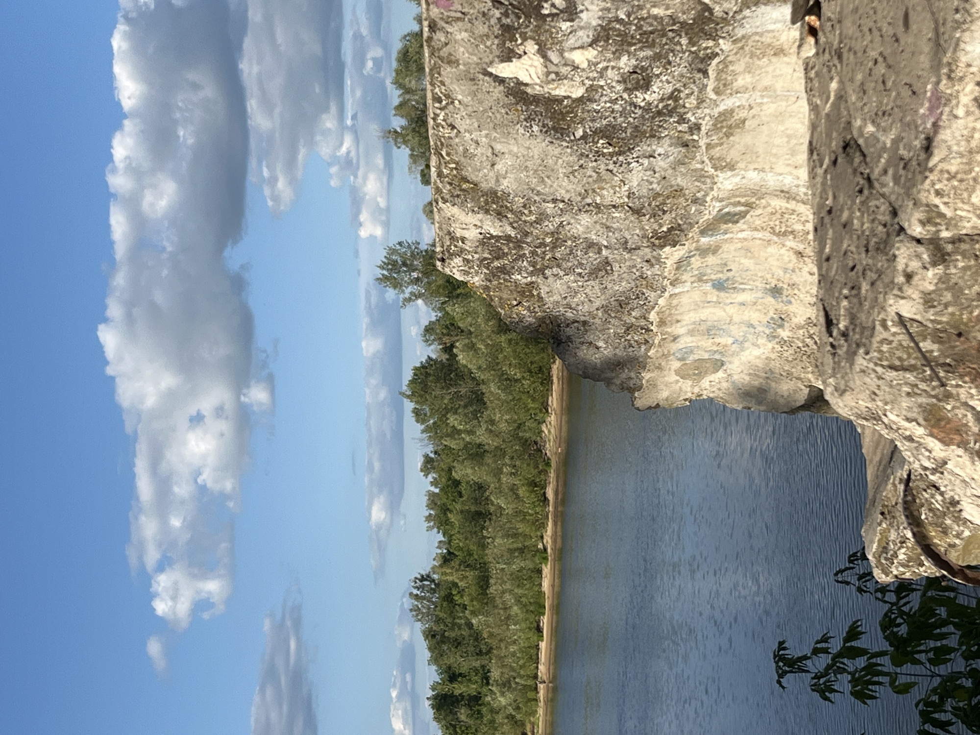 The image depicts a natural scene with a body of water in the foreground and a wooded area in the background. A large, weathered concrete structure, possibly a remnant of industrial infrastructure, occupies the right side of the frame. The water, appearing calm, reflects the blue sky dotted with white cumulus clouds. Dense green foliage lines the opposite bank of the water, suggesting a lush natural environment. The concrete structure shows signs of age and weathering, with patches of moss and what appears to be faded graffiti. Small green plants are visible in the lower left corner, growing near the edge of the concrete. The overall impression is one of a somewhat neglected, but scenic, outdoor location.