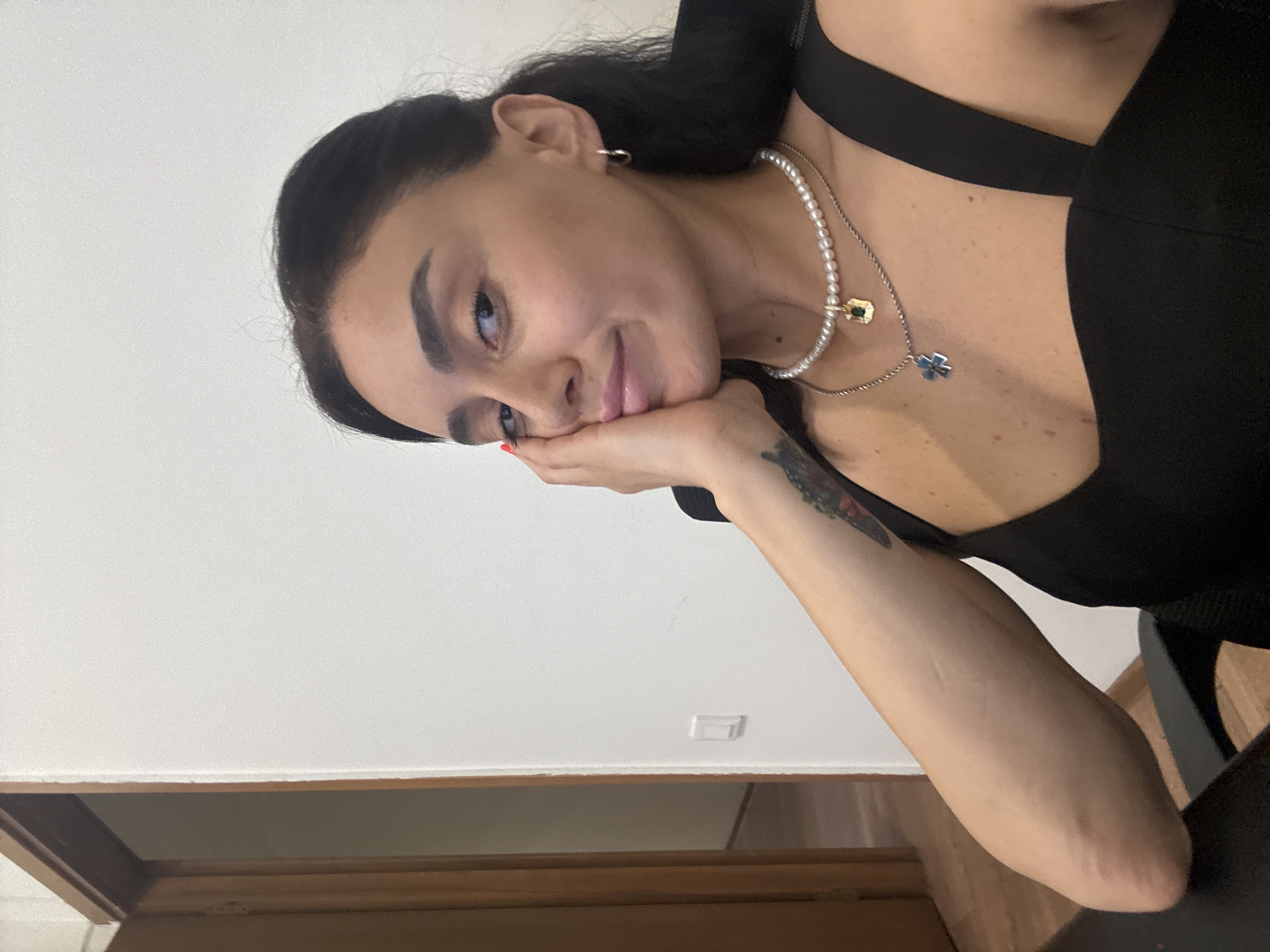 The image shows a close-up, selfie-style shot of a woman with dark hair and fair skin. She is wearing a black top with thick straps and two necklaces: one with pearls and a yellow pendant, and another with a silver chain and a blue cross pendant. She has a tattoo on her left forearm. Her right hand is resting on her cheek, and she is looking towards the camera with a slight smile. The background is a plain white wall with a doorway visible on the left. The scene appears to be indoors, in an office or residential setting. There is no indication of the location being in Tbilisi, Georgia.