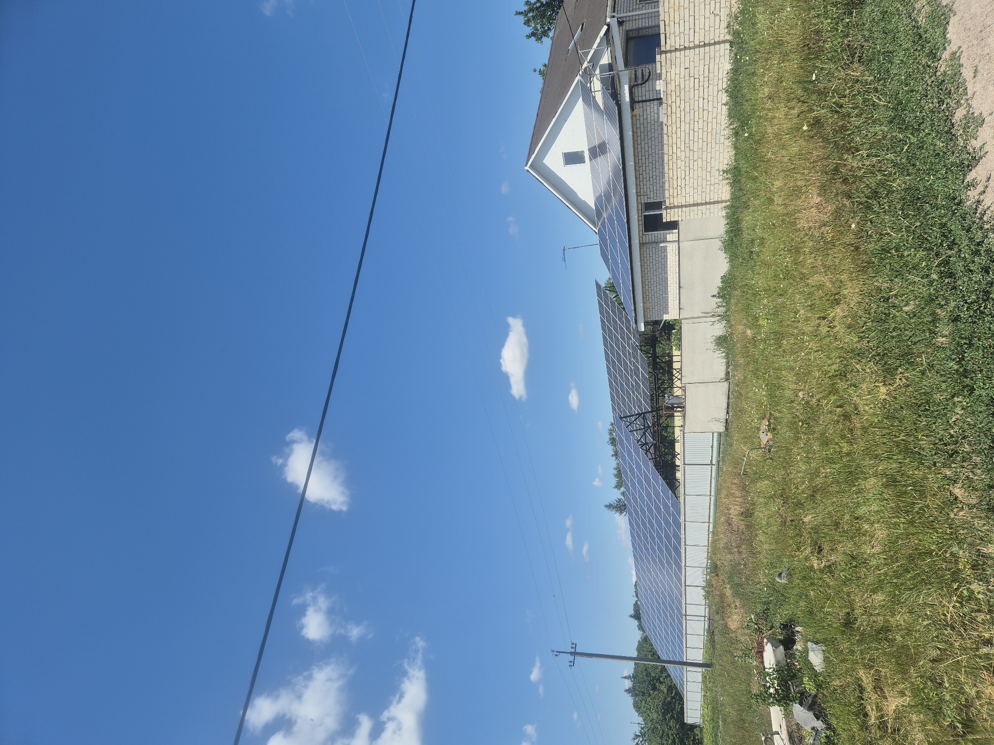 In Sozonivka, Ukraine, a sunlit scene unfolds with a clear blue sky dotted with scattered white clouds. In the foreground, a patch of tall, green grass with some dried stalks and scattered wildflowers occupies the lower portion of the image. A dirt path or road is visible at the very bottom edge.
Dominating the middle ground is a large array of solar panels, mounted on a dark metal frame. This array stretches horizontally across the frame, extending from behind a light-colored, chain-link fence that runs parallel to the solar panels. Adjacent to the solar array, on the right side of the image, is a white brick building with a brown roof, also fitted with solar panels. Power lines and a utility pole are visible to the left of the solar array, with two parallel wires stretching across the upper part of the image.
The background features a dense line of green trees. The overall environment appears to be a rural or semi-rural setting, likely a property with a residential building and a significant solar energy installation. There are no people or animals visible in the frame.