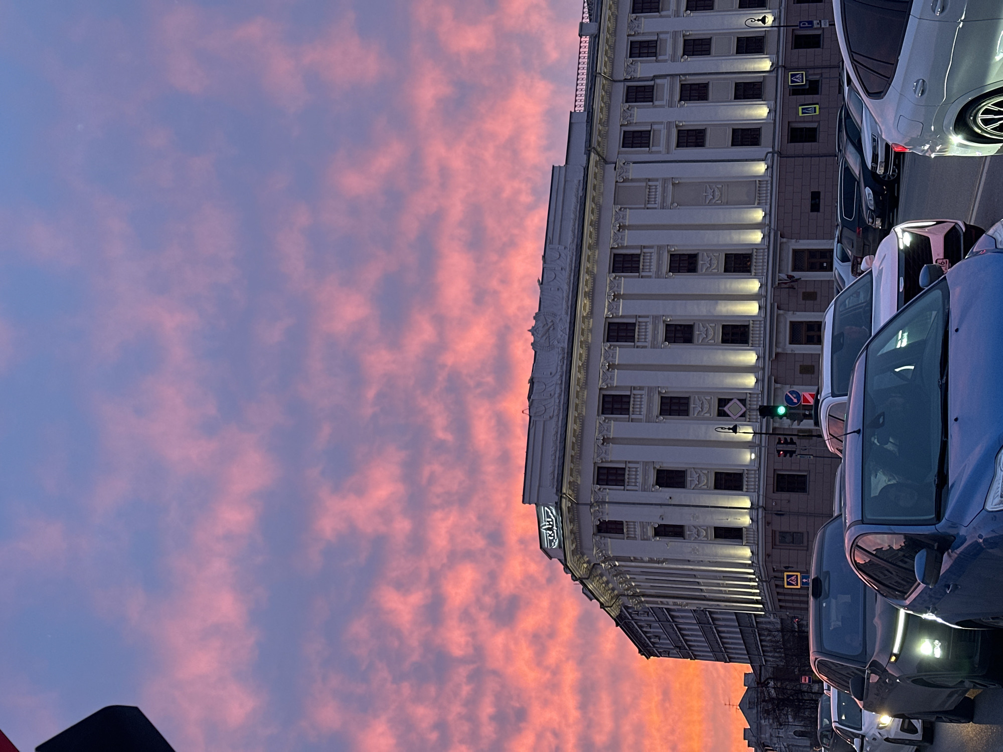 The image depicts a street scene in Kazan, Russia at dusk. A tall, ornate, light-colored building occupies the right side of the frame, adjacent to several cars lined up on the street. The sky overhead is filled with clouds that are illuminated with a pink and orange hue, indicating sunset. The building features multiple windows with visible lighting, and a prominent sign with a logo. Traffic lights are visible, with green and red colors illuminated. Several cars are parked on the street.