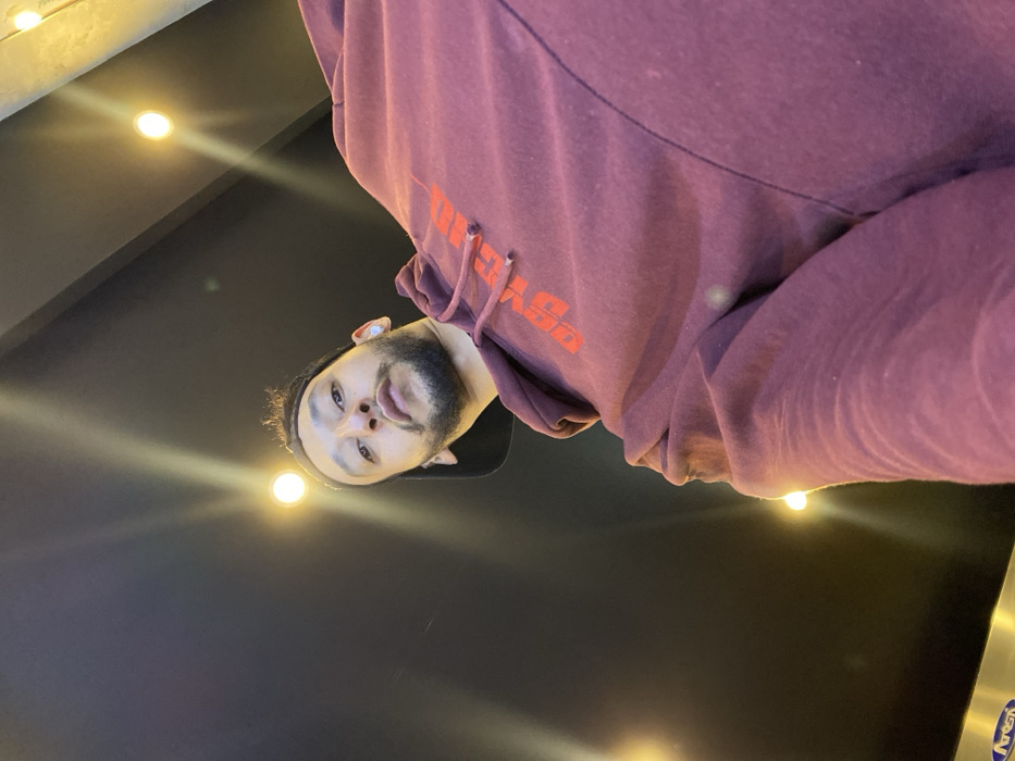 A man looks down at the camera, wearing a maroon hoodie with orange text. He has a dark beard and a light complexion. The background is dark with several bright, circular lights arranged across the top. The camera angle is low, looking up at the man.