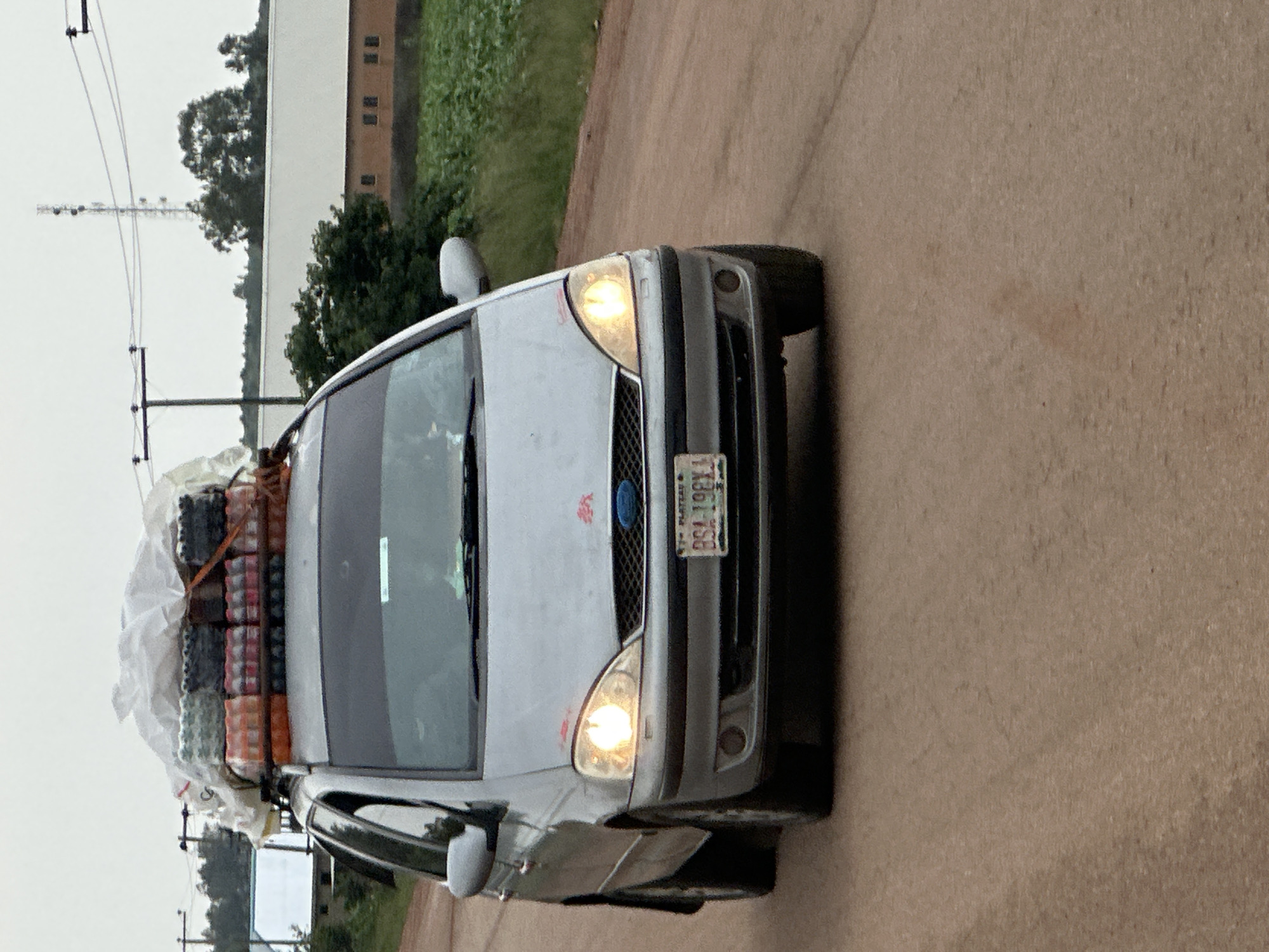 A silver Ford Galaxy minivan is traveling on a dirt road. The minivan's roof rack is loaded with many bottles of drinks, likely soda, covered with white plastic sheeting and secured with straps. The front of the minivan has its headlights on. A license plate on the front of the minivan reads "BSA 198X1 PLATEAU". The environment appears to be rural, with a field of crops on the right side of the road and trees and buildings in the background. There is a power line tower visible in the distance. The sky is overcast.
