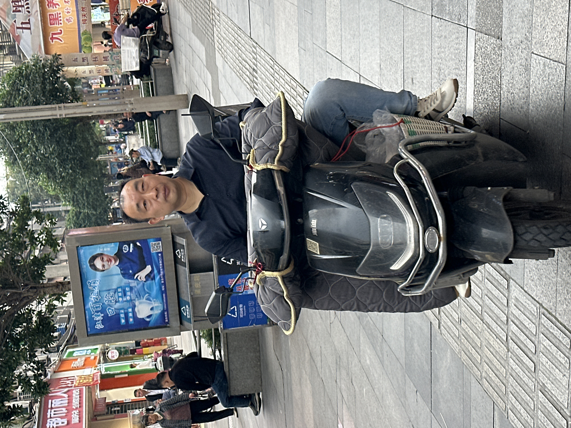A man in a navy shirt and blue jeans is sitting on a black motorcycle on a city street in Jiangjin, China. He is looking directly at the camera. The motorcycle has quilted hand covers and a chrome front bar. The sidewalk is gray and textured. Behind the man are pedestrians and storefronts with Chinese characters. A large digital advertising display features a woman's face and Chinese writing.