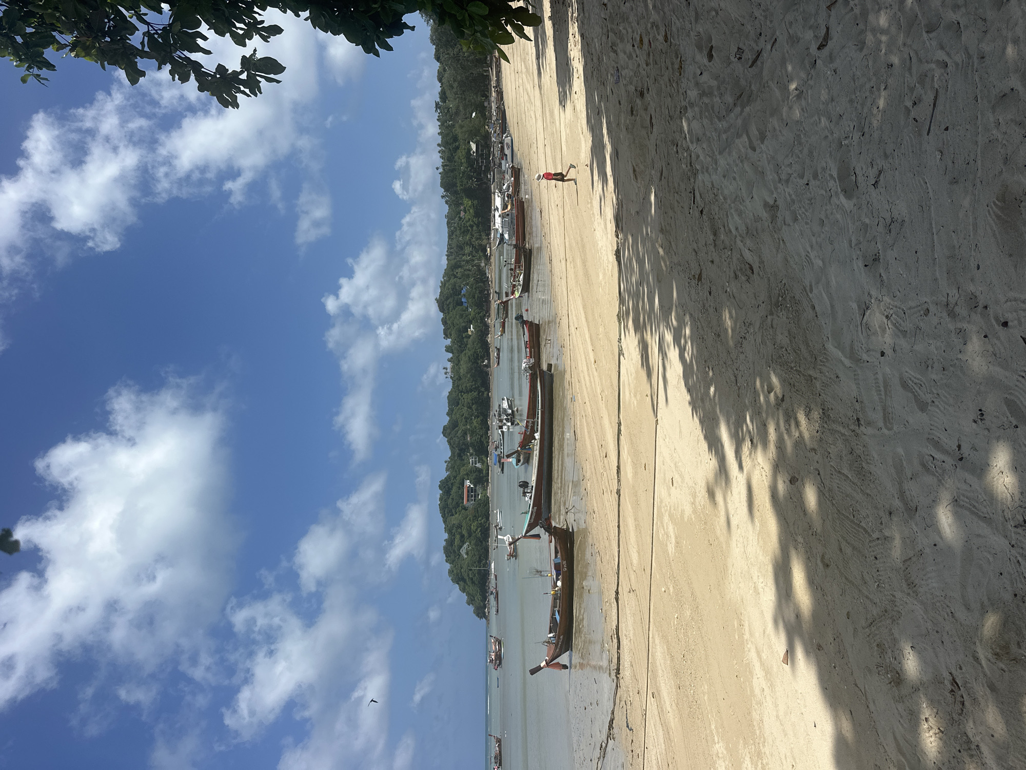 A beach scene at Rawai, Thailand, under a bright blue sky with scattered white clouds. The foreground is a sandy beach with shadows from trees cast across it. In the mid-ground, a line of traditional Thai longtail boats are moored in shallow water, some resting on the sand as the tide is out. Further out in the calm sea, more boats are visible, and in the distance, a lush green, tree-covered hill rises from the water. A single person wearing a red shirt and dark shorts is walking along the beach. The overall atmosphere is one of a peaceful tropical day.