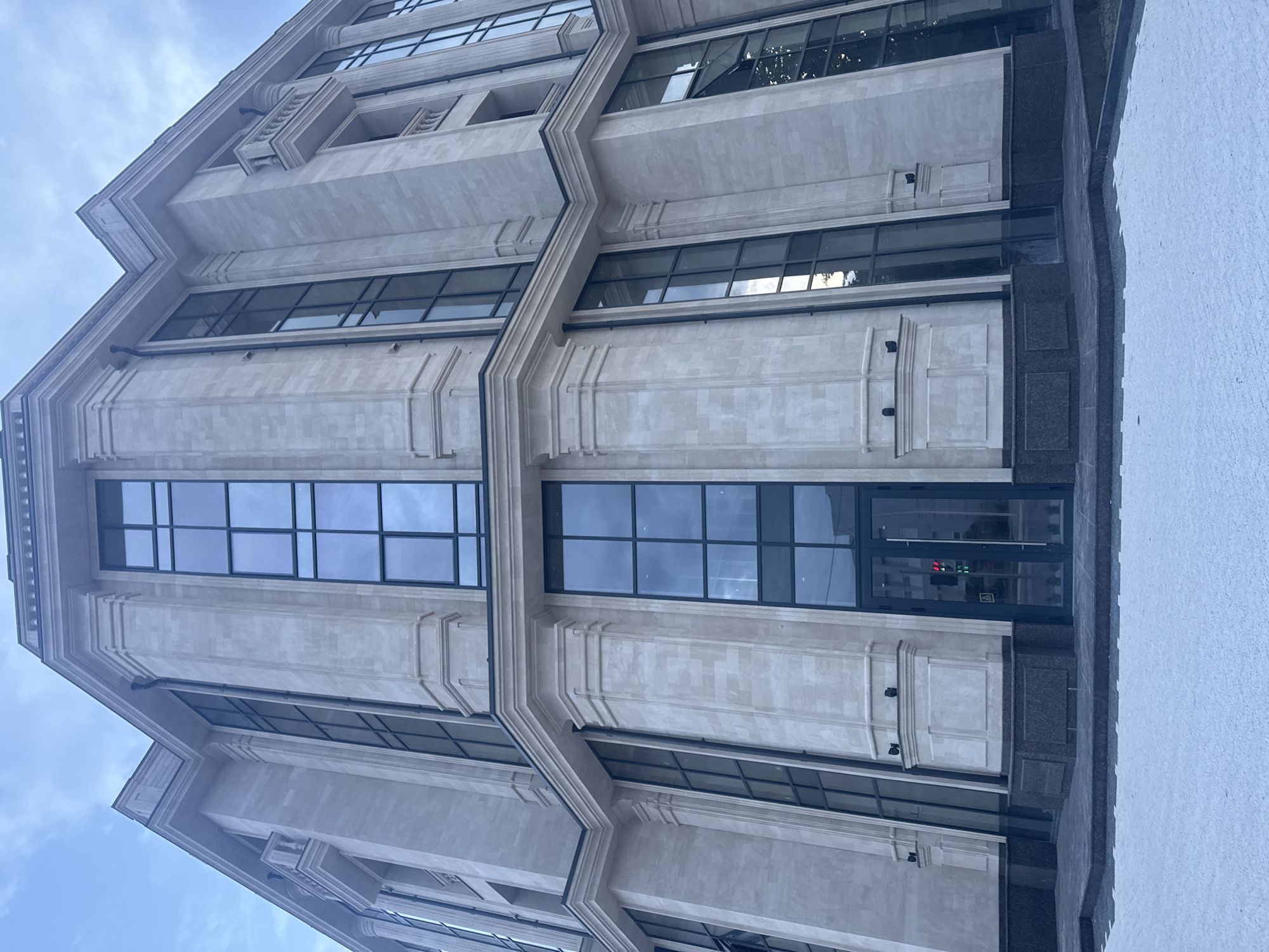 A low-angle, wide shot captures the facade of a modern, multi-story building constructed with light-colored stone. The building features numerous tall, dark-framed windows and decorative architectural elements, including columns and cornices. A double glass door entrance is visible at the base of the building. The sky above is overcast and appears to be either dawn or dusk. The ground in front of the building is paved with dark stone and a lighter, textured material. No people are visible in the image.