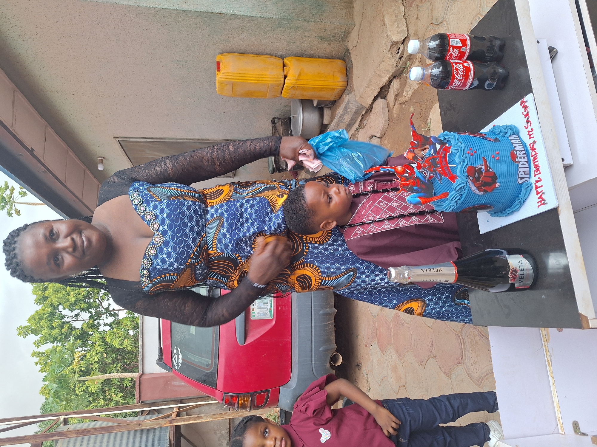 This daylight outdoor photograph, taken in Sabon Tasha, Nigeria, captures a small birthday celebration featuring three individuals.
In the center, a woman with dark skin and braided hair smiles warmly at the camera. She is wearing a blue dress with a vibrant geometric pattern in blue and orange, and black lace long sleeves. She holds a light blue plastic bag containing an indistinct item in her right hand. Standing in front of her is a young boy, likely the birthday celebrant, wearing a maroon patterned shirt over a plain maroon one, looking towards the right. To the lower right of the frame, another young girl with dark skin, dressed in a maroon top with puffy sleeves and blue jeans, gazes directly at the camera.
A blue "Spiderman" themed birthday cake is prominently displayed on a dark-colored table to the right. The cake is decorated with a Spiderman figure and "SPIDERMAN" written on its side. It rests on a white cake board bearing red icing text that reads "HAPPY BIRTHDAY KI. SUCCESS". Beside the cake are two plastic bottles of "Coca-Cola ORIGINAL TASTE" and a bottle of "VELETA" sparkling drink.
The background reveals an outdoor setting. A red car, appearing to be a hatchback or SUV, is parked behind the woman, with "NIGERIA" partially visible on its license plate holder. Beyond the car, lush green trees and foliage are visible under a bright sky. To the left, near a building, are two yellow plastic jerrycans and a metal pot. The ground is a mix of paved and dirt surfaces. The scene conveys a joyful and informal gathering, likely celebrating the young boy's birthday.