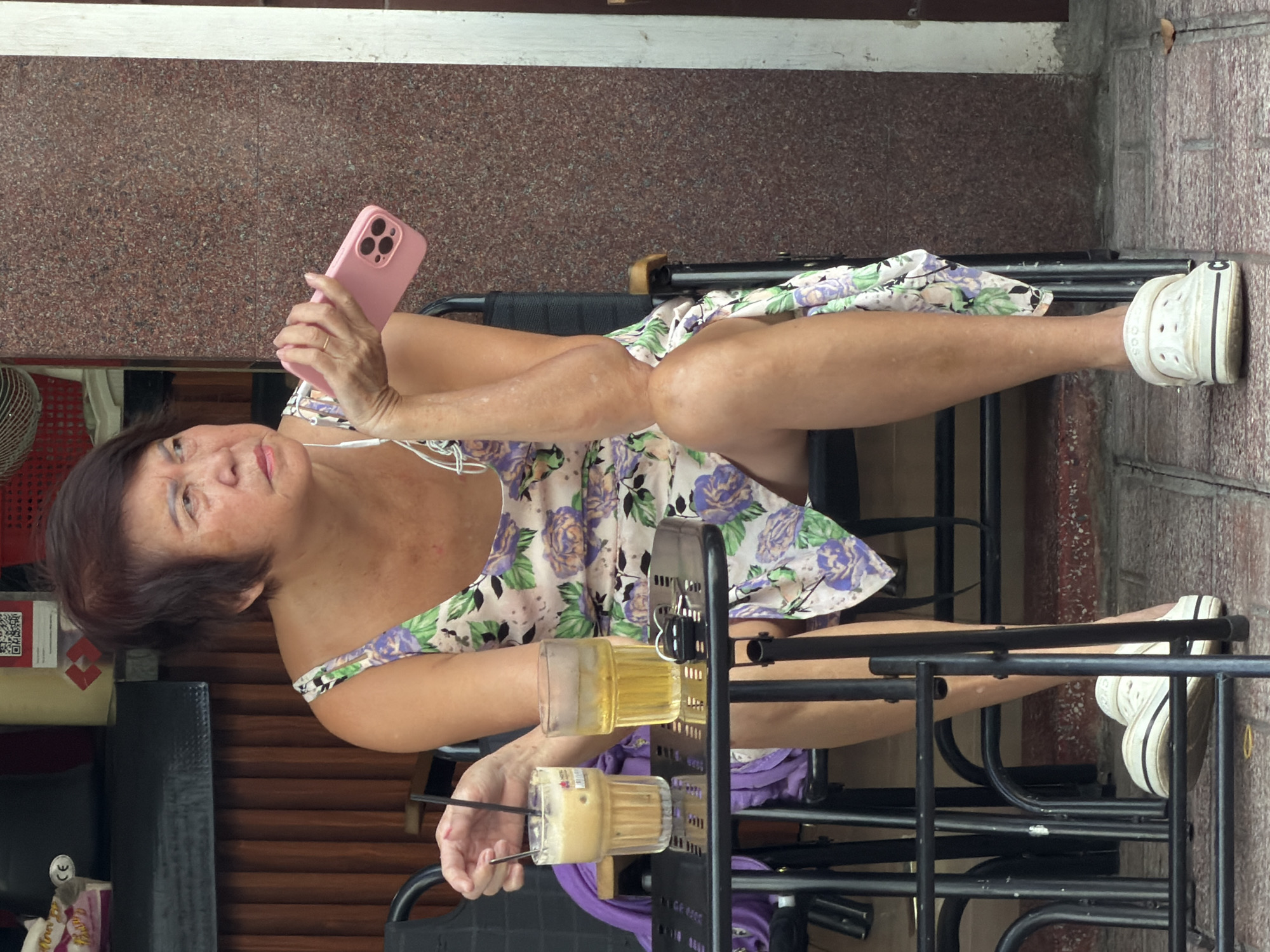 A Vietnamese woman sits at a small black table outside a building with a textured brown facade and wooden slatted siding. She is wearing a light-colored, floral-print dress with purple and green flowers and white clogs. She holds a pink smartphone in her right hand and has earbuds in her ears. On the table are two drinks: one tall glass filled with a light brown iced coffee and another glass with a yellow liquid and ice. A set of keys is also on the table. The background shows a brick-patterned sidewalk and part of a doorway. The scene appears to be a street-level cafe or outdoor seating area in Nha Trang, Vietnam.