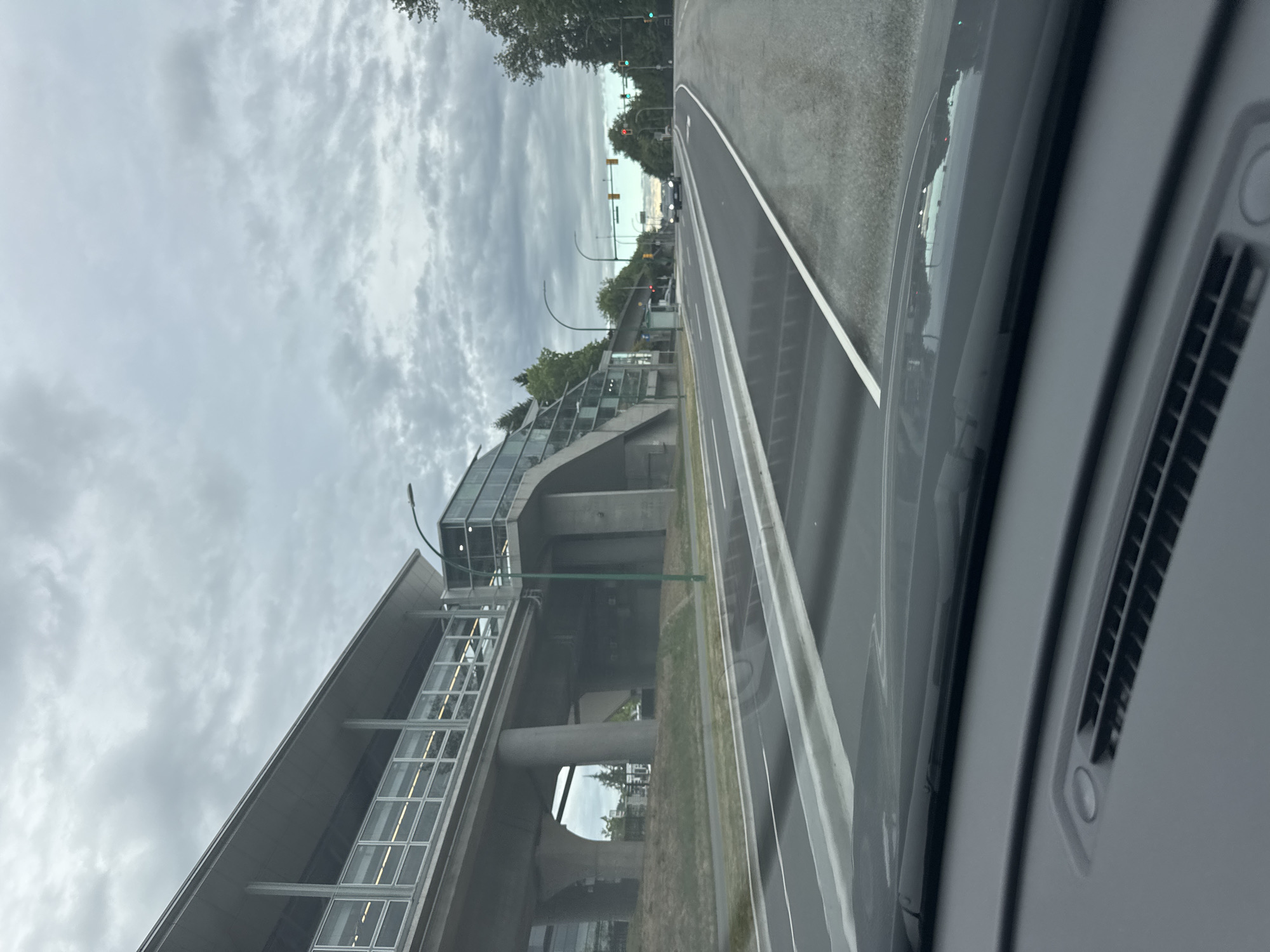 The image captures a view from inside a vehicle looking forward on a cloudy day. A modern elevated transit station with a glass facade runs along the left side of the road. The station is supported by concrete pillars. A street lamp with a green pole is visible near the station. The road ahead has multiple lanes and is wet, reflecting the overcast sky. Several cars are on the road, and traffic lights are visible in the distance, with some showing green and one showing red. Trees line the right side of the road, and more of the transit station structure is visible further down. The foreground shows the dashboard of the vehicle. This scene is identifiable as the Edmonds SkyTrain Station in Burnaby, Canada.
