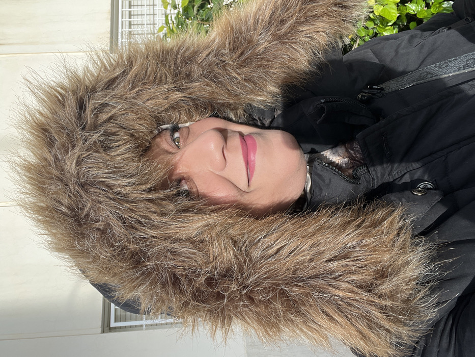A female subject, appearing middle-aged, is depicted in a close-up, chest-up outdoor portrait, set in Meknes, Morocco. She is wearing a dark (likely black) quilted jacket with a prominent, voluminous, light brown faux fur-trimmed hood that frames her face. Her head is slightly tilted, and she exhibits a closed-mouth smile. Visible facial features include dark eyes with mascara, defined eyebrows, and pink lipstick. A light-colored, possibly patterned, garment is visible beneath the jacket's collar. Strong sunlight illuminates the scene, casting shadows on the subject's right side and jacket. The background features a light-colored architectural structure with visible horizontal window elements on the left, and vibrant green foliage on the right.