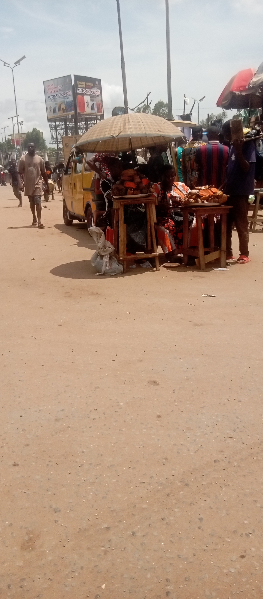 In a street scene in Onitsha, Nigeria, vendors hawk their wares from makeshift stalls set up on wooden tables under a large straw umbrella. A woman, dressed in an orange and floral patterned dress, sits near the tables, possibly selling goods. The tables are laden with what appears to be produce or cooked food. People mill around the stalls, some seemingly browsing. Adjacent to the stalls, a yellow commercial bus is parked. In the background, a billboard advertises AUTO-SHECK OIL. The ground is a dusty brown color. A man walks toward the camera on the left side of the frame, and other figures can be seen in the distance.