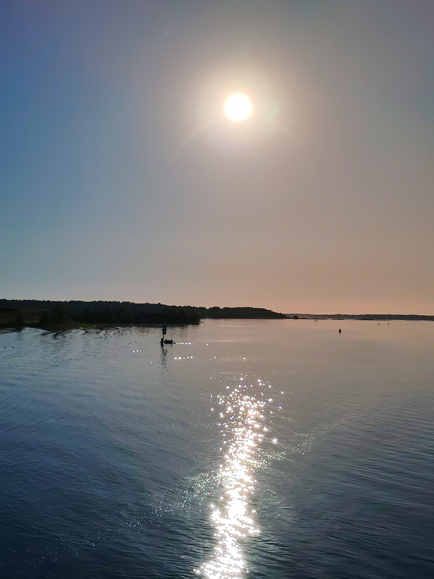 This image depicts a serene maritime landscape, likely situated in the city of Mys, Russia. The scene is dominated by a wide expanse of calm water under a brightly illuminated sky. The upper sky transitions from a soft blue on the left to a warm, hazy off-white and pale orange hue on the right, where a brilliant, white, circular sun is centrally positioned, casting intense glare and visible light rays. Scattered glints, possibly atmospheric dust, are noticeable around the sun.
The water surface is dark blue-gray on the left, showing subtle wave patterns. A prominent, shimmering path of sunlight extends from the sun towards the viewer, creating numerous sparkling reflections across the water. On the left side of this reflection path, a dark navigation buoy stands upright. To the left of the buoy, a small, dark boat, occupied by one or two individuals, is present, generating a minor wake.
The left shoreline features a low-lying landmass densely covered with dark trees or vegetation, extending into the mid-ground. Further in the distance, a faint, linear landmass is visible along the horizon on the right. Several other small, dark buoys or markers are sparsely distributed across the distant water. The overall lighting suggests a bright, clear daytime environment.
