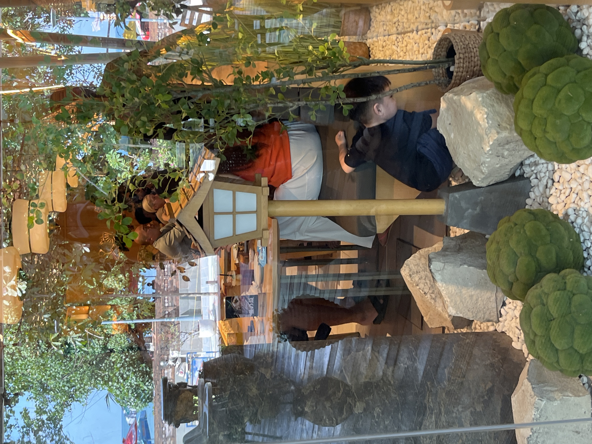 This image, taken in Kuta, Indonesia, appears to be an indoor scene viewed through a glass pane, possibly a shop window or a restaurant. The reflection on the glass distorts the view and creates a layered effect.
The primary subjects are two young boys. One boy, on the right, is wearing a dark blue or black outfit and is looking towards the left. He is positioned near a large, light-colored rock and some decorative green moss balls. The other boy is more obscured, seen from behind, wearing a dark shirt. He appears to be sitting or crouching. Two adults are also partially visible. One is wearing an orange top and light-colored pants, and the other is wearing a grey top.
The setting seems to be a retail space or a themed restaurant, possibly with a Japanese or natural aesthetic. There are plants, both real and artificial, and decorative elements like stones, white pebbles, and a basket. A small wooden structure with a lamp-like feature is also present, resembling a miniature traditional Japanese house. The reflections show trees, buildings, and a blue sky, suggesting the scene is viewed from inside looking out, or vice-versa, with significant reflection.
The lighting suggests it is daytime, with natural light illuminating the scene. The overall mood is somewhat serene, with the presence of children and the natural elements. There is no visible text in the image.
