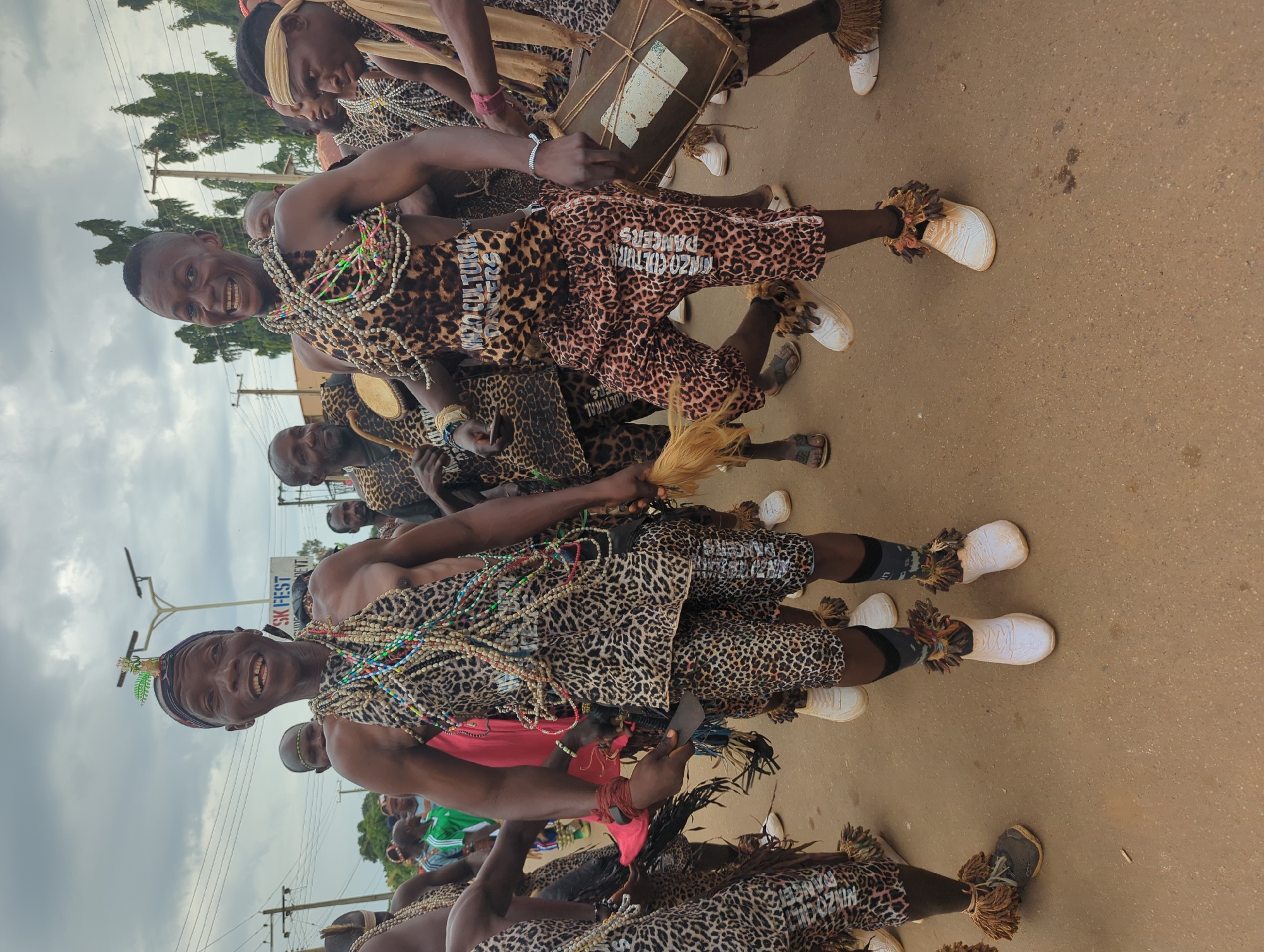 A group of men, identified as "Nzo Cultural Dancers" by the lettering on their attire, are performing in Maigizo, Nigeria. They are dressed in matching leopard-print outfits consisting of shorts and tops, adorned with multiple strands of colorful beads. The dancers are also wearing white sneakers with fringed ankle decorations. Some are playing drums, while others are holding feathered objects. The street scene shows a cloudy sky, utility poles, and other onlookers in the background.