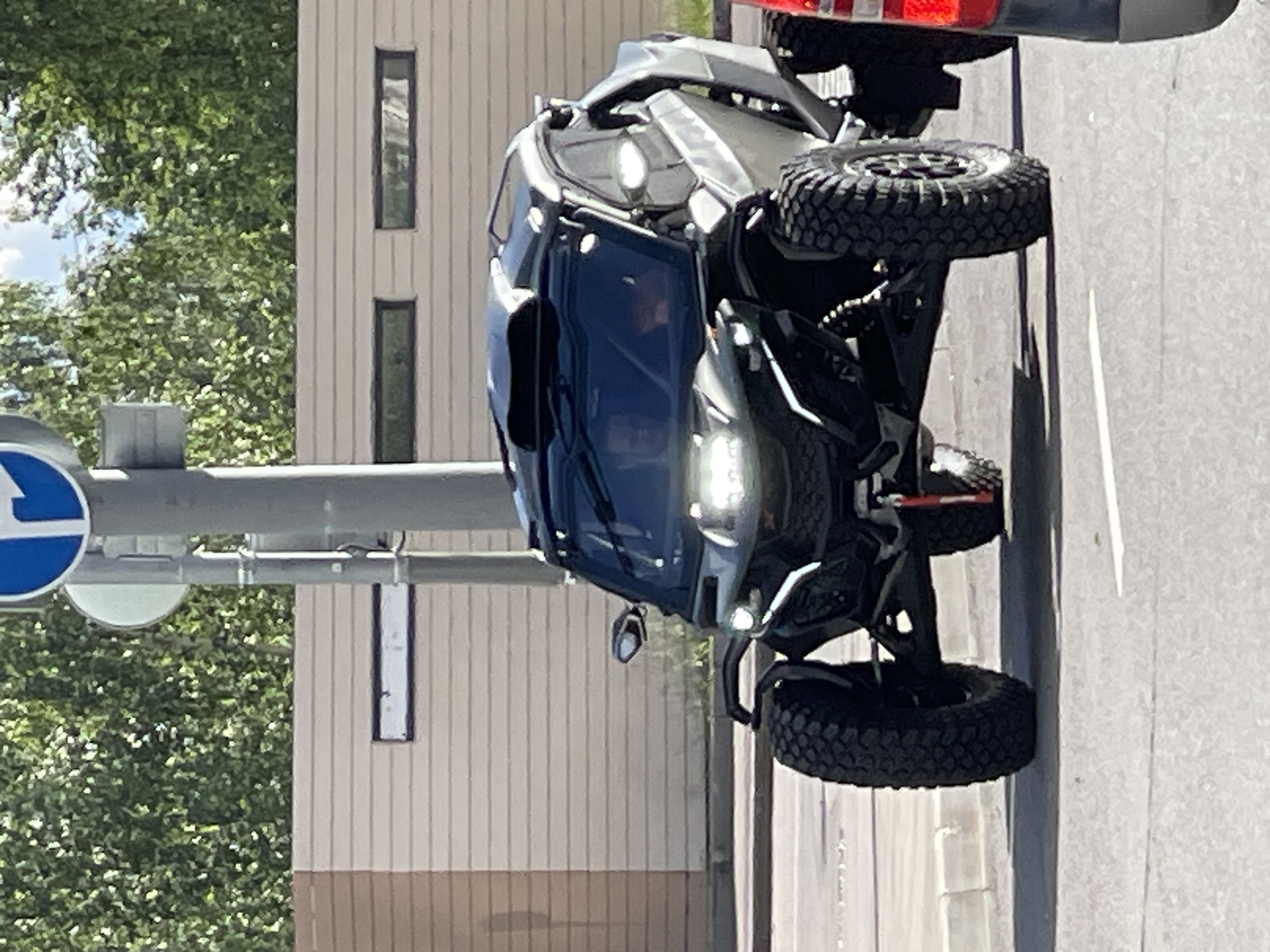 The image shows a black all-terrain vehicle (ATV) parked on an asphalt road. The ATV has large, knobby tires and appears to be a sporty, open-cockpit model. The sun is shining, indicating it is daytime, and the weather seems to be clear.
In the background, there is a light-colored building with vertical siding and several rectangular windows. Adjacent to the building is a dense green tree, suggesting an urban or suburban setting with some landscaping. A gray pole with a blue and white traffic sign is visible on the left side of the frame. The overall scene suggests a sunny day in a modern environment. There are no people clearly visible in the image, and no text is discernible. The image is rotated 90 degrees counterclockwise.
