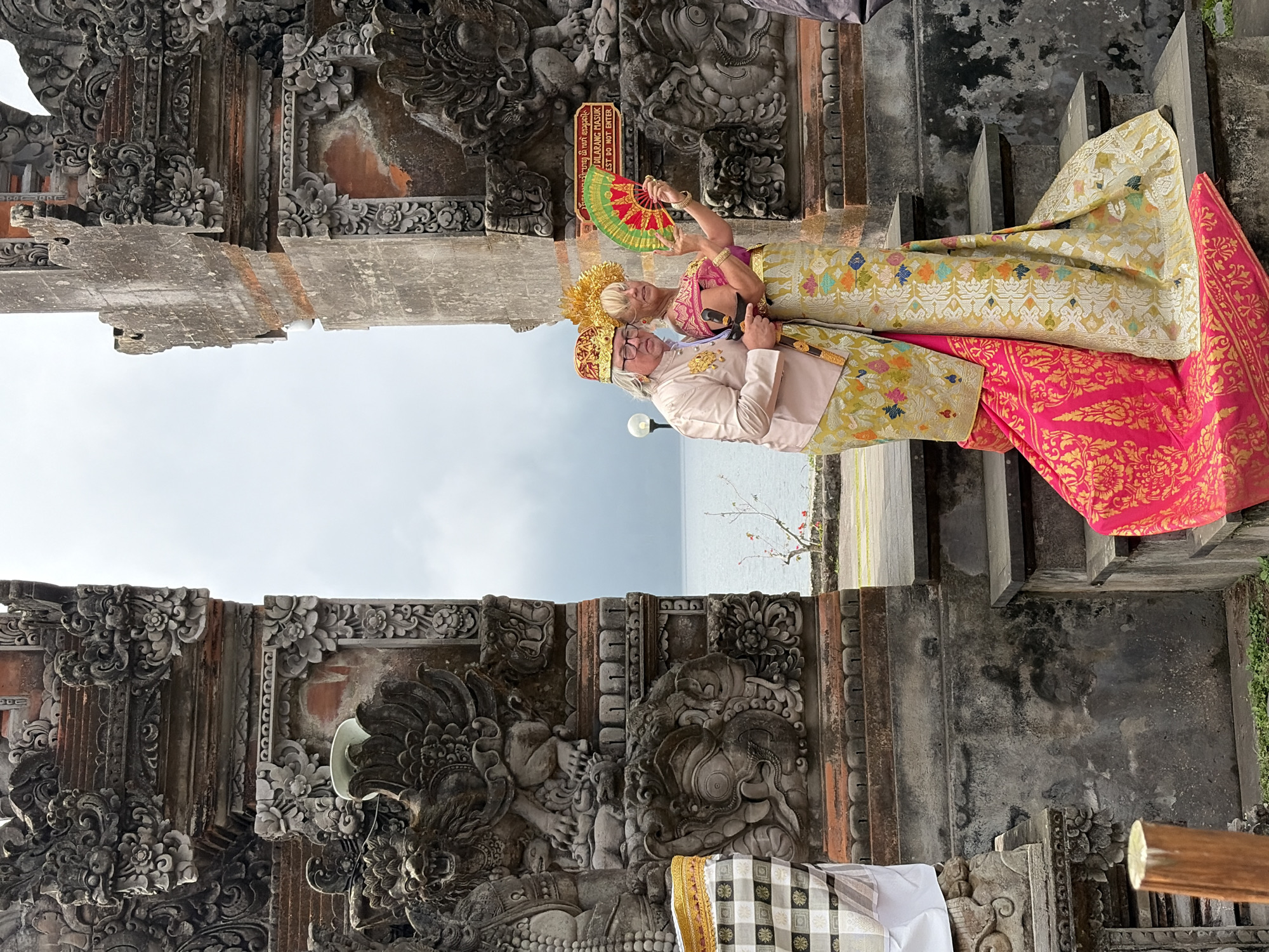 The image shows two people, a man and a woman, dressed in traditional Balinese attire standing on stone steps in front of a weathered stone temple structure. The man is wearing a pale pink tunic, a gold and red patterned hat, and glasses. The woman is wearing a gold headdress, a pink and gold top, and a long skirt with intricate patterns in gold, green, pink, and blue. She is holding a red and green fan and has her left arm around the man's waist. The temple structure is elaborately carved with mythical figures and floral motifs. In the background, the ocean is visible under an overcast sky, with a few bare tree branches with small red flowers in the foreground. A sign with Indonesian text is visible on the temple structure behind the woman. The overall scene suggests a cultural or ceremonial event at a historical site. The location is identified as Baturiti, Indonesia.