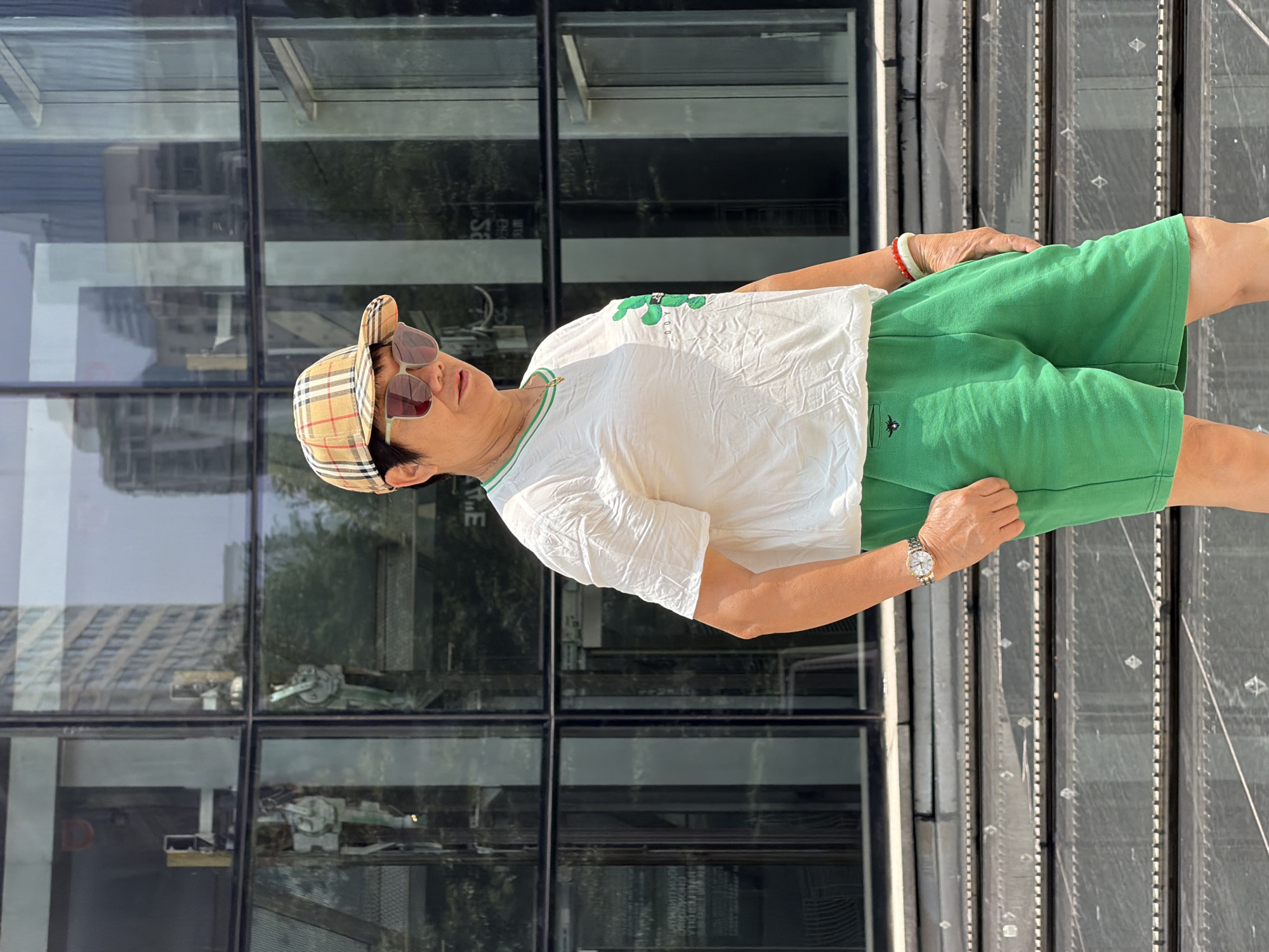 An adult with short dark hair, wearing sunglasses and a plaid baseball cap, stands in front of a building with a glass facade. They are wearing a white t-shirt with a green design on the chest and bright green shorts. A silver watch is visible on their left wrist, and red and jade bracelets are on their right wrist. The background shows reflections of buildings in the glass facade. The person is in Yuzhong District, China.