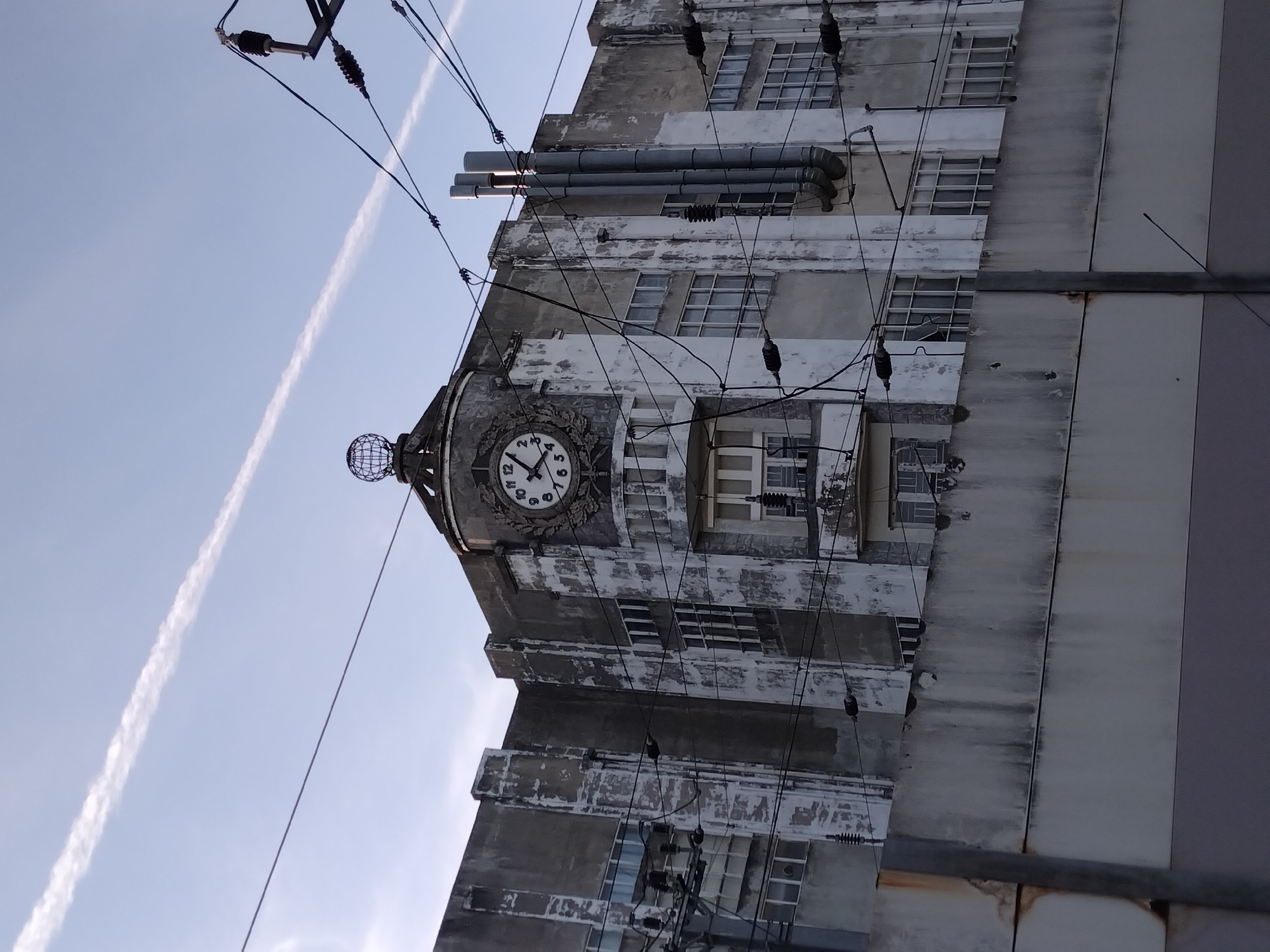 The image shows a close-up, low-angle view of the facade of an old, weathered building, likely an industrial or administrative structure, in Vila Nova de Famalicão, Portugal. The building has a distinct clock tower at its apex, topped with a globe-like ornament. The clock face, with Roman numerals, is visible, with the hands indicating approximately 12:45. The building's concrete exterior is peeling, revealing patches of white underneath the grey, suggesting age and perhaps neglect. Multiple power lines and electrical insulators crisscross the frame, obscuring parts of the building and indicating the presence of an active utility infrastructure. Several tall, metallic pipes, possibly ventilation or exhaust systems, are attached to the side of the building. The sky above is a pale blue with a prominent contrail stretching diagonally across it. In the foreground, a gray concrete wall runs horizontally, further framing the building. No people are visible in the scene.