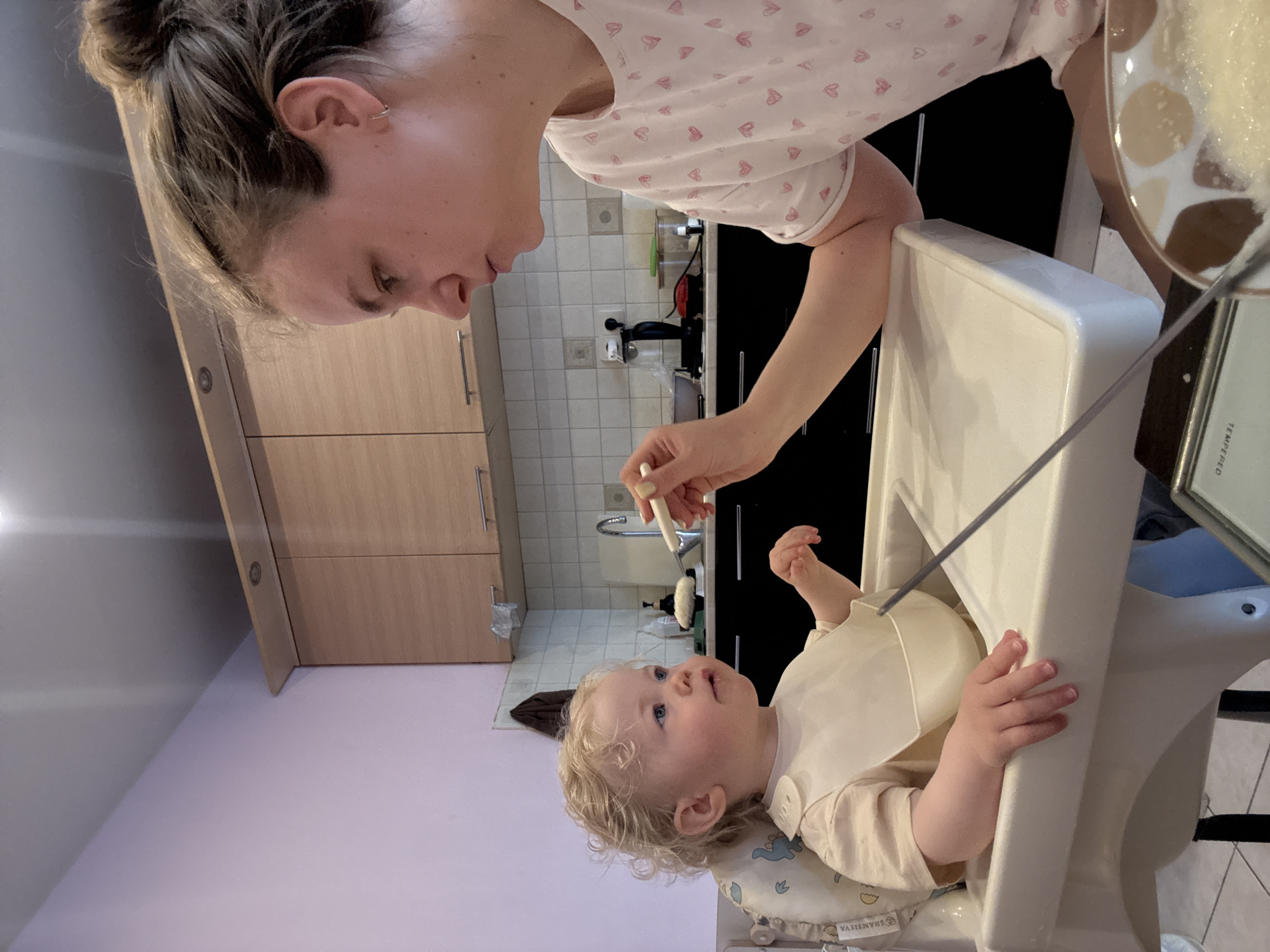 A mother is feeding her baby, who is sitting in a high chair. The mother is holding a spoon and looking at the baby. The baby is looking up at the mother. The background shows a kitchen with cabinets, a sink, and a wall. The city and region are Unknown.