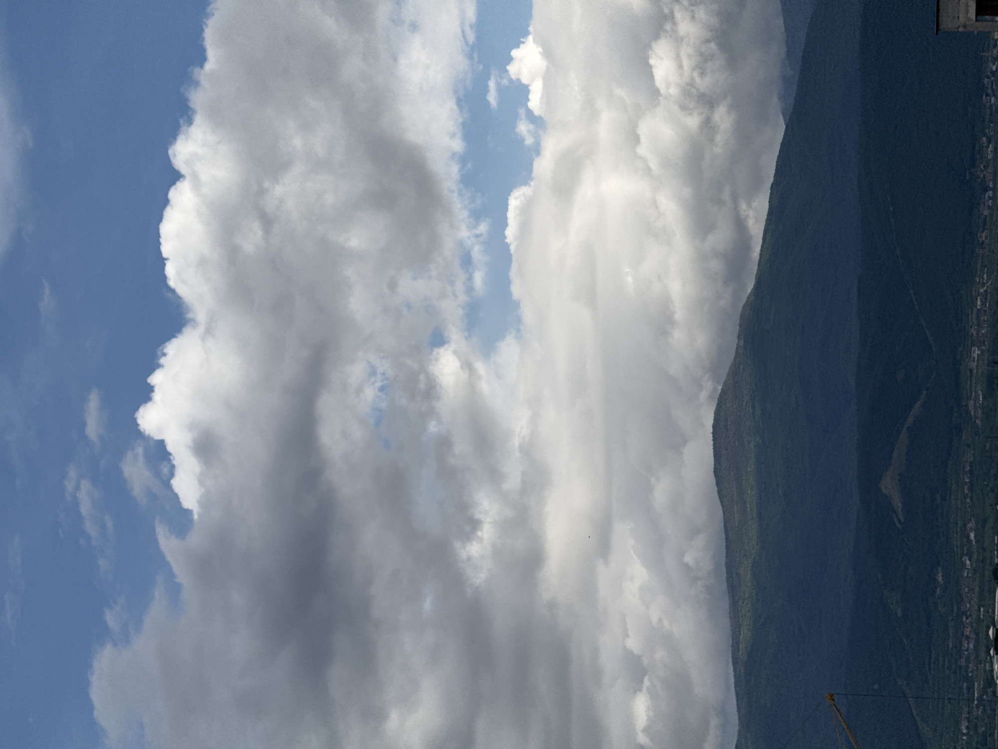 The image depicts a landscape with a large mountain range on the right side, its slopes covered in lush greenery. A city or town is nestled at the base of the mountain, with buildings and roads visible. The sky is mostly filled with thick, voluminous clouds in varying shades of white and gray, leaving only narrow strips of blue visible. The photograph appears to have been taken during daylight. The bottom portion of a yellow crane is visible. The image is taken in Plovdiv, Bulgaria.