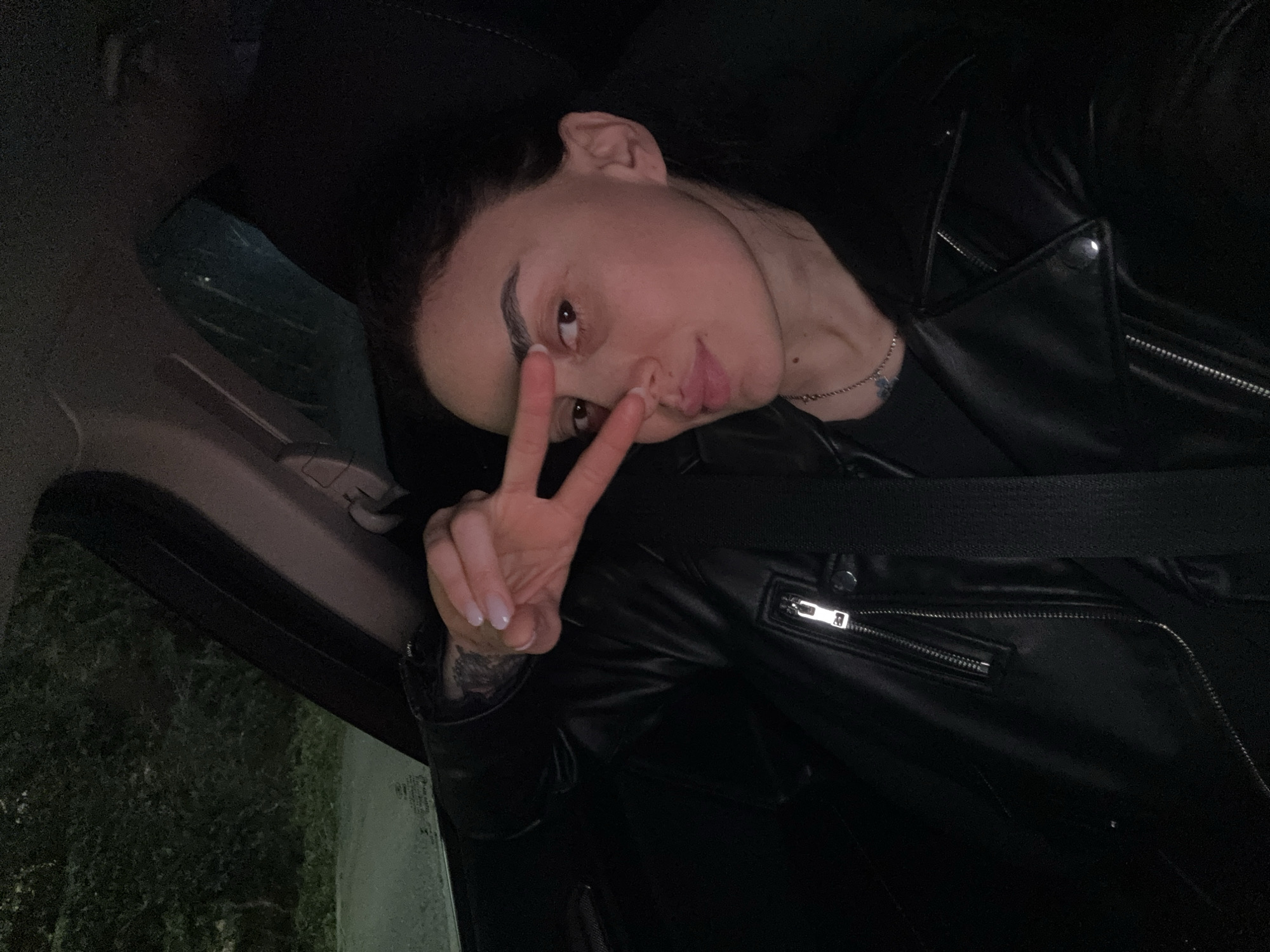 A young woman is prominently featured in a diagonal portrait, likely a selfie, taken inside a car in Tbilisi, Georgia. She is positioned towards the center-right of the frame, making a peace sign (V-sign) with her right hand held up to her face. She looks directly at the camera with a slight pout or smirk.
She wears a black leather jacket with silver zippers and snaps. A silver necklace with a small, light-colored charm is visible around her neck. A seatbelt crosses over her chest, indicating she is a passenger in the vehicle. Her fingernails are painted with a light-colored polish.
The car's interior is partially visible, including parts of the headrest, ceiling, and the side window. The background outside the window is dark and indistinct, suggesting it is nighttime or heavily overcast. There appear to be dense trees or bushes outside, and possible moisture or reflections on the car window. The overall lighting is dim, casting shadows and highlighting her features against the dark surroundings. No text is visible in the image.