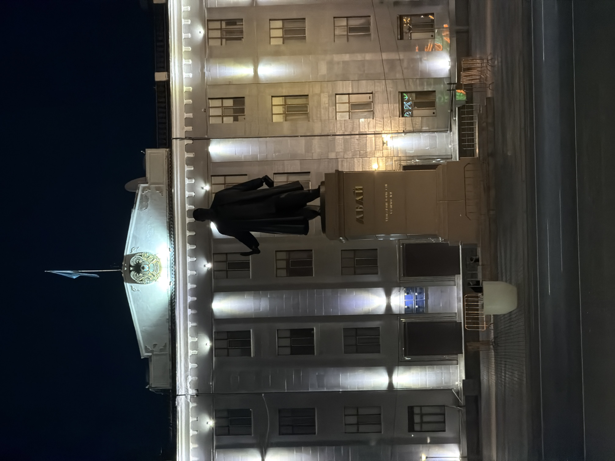 In the city of Pavlodar, Kazakhstan, at night, a statue of a man in a coat stands on a pedestal in front of a large, illuminated building. The building is several stories high with many windows and decorative architectural elements. A flag flies from the top of the building. The area in front of the building appears to be a public space with a paved walkway and some temporary barriers. The lighting suggests it is evening or night. The statue is the central focus of the foreground, with the building serving as a backdrop.