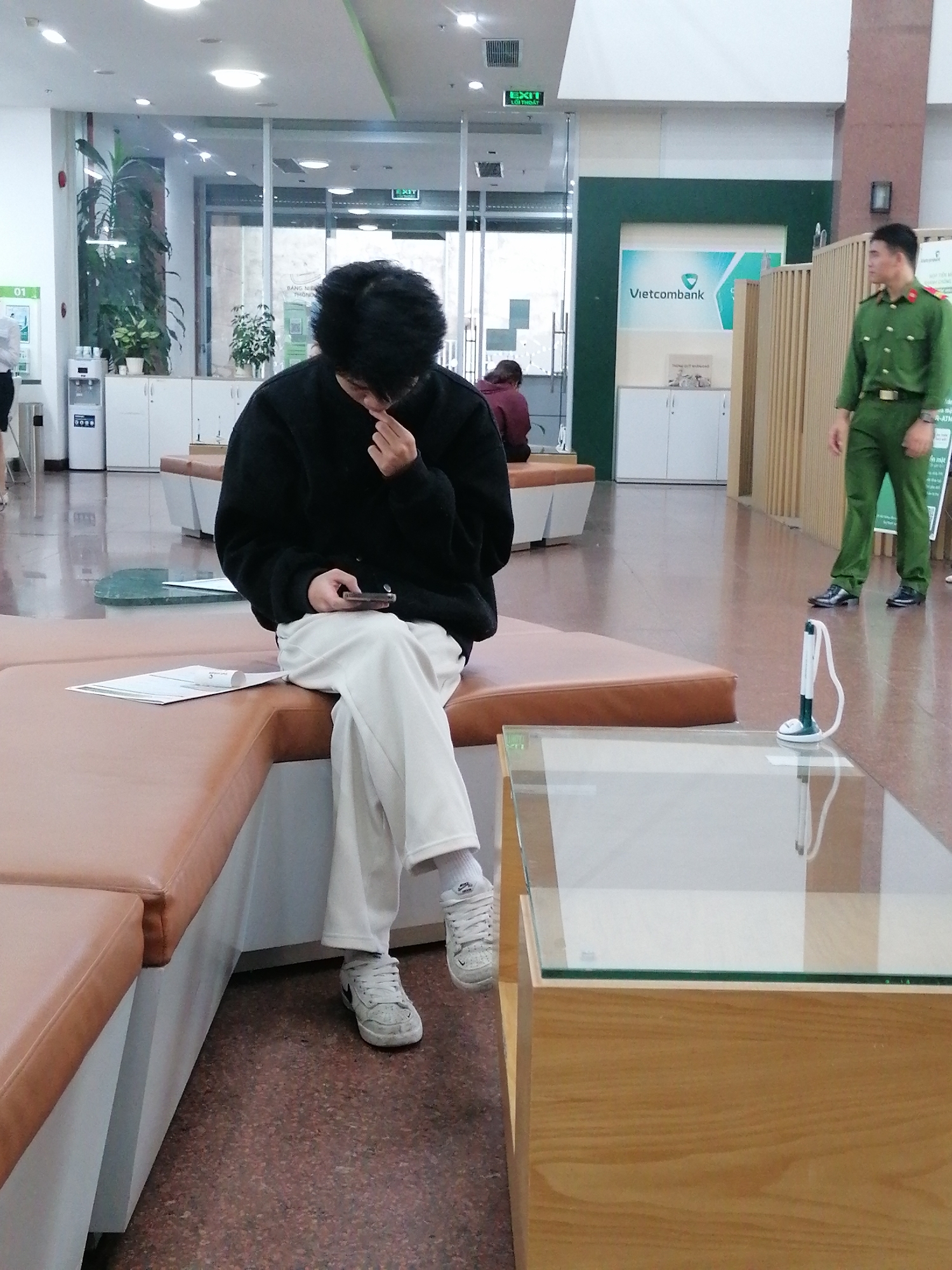 In a brightly lit interior space, possibly a bank lobby in Da Lat, Vietnam, a young person with dark, tousled hair is seated on a long, brown, leather-upholstered bench. They are wearing a black, oversized sweater and loose-fitting, cream-colored pants, with white athletic shoes. The person is looking down at a smartphone held in their hands, touching their face with their right hand as if contemplating or tired. To the left of the bench, a water dispenser and white cabinets are visible against a light-colored wall.
Further into the scene, behind a partition with vertical wooden slats, a uniformed man in a dark green outfit stands facing to the left. He has dark hair and appears to be on duty, possibly a security guard. The wall behind him features a large green and white logo for "Vietcombank," a Vietnamese commercial bank. In the background, a reflection on the polished floor shows ceiling lights and office-like structures. The environment suggests a public service area, likely a financial institution, with elements like an "EXIT" sign and informational posters visible on the walls. A low, glass-topped coffee table with a light wood base is positioned in the foreground, reflecting the surroundings. The overall atmosphere is calm and orderly.