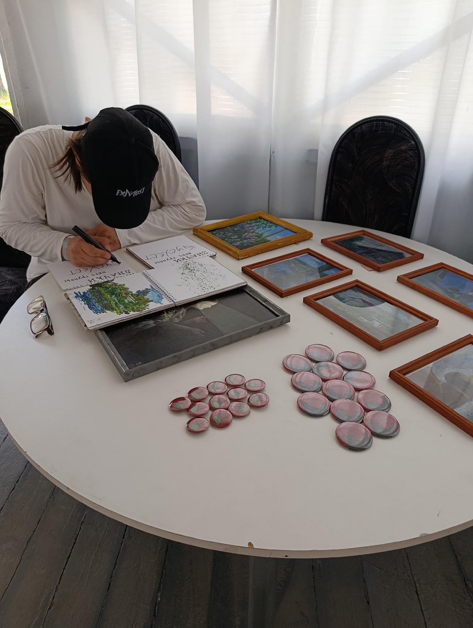 A person wearing a white long-sleeved shirt and a black baseball cap is sitting at a round white table, drawing in an open notebook. The notebook lies on top of a dark gray rectangular object. To the left of the notebook are a pair of eyeglasses. Several framed pictures with wooden frames are arranged on the table, some containing artwork. In the foreground, to the right, are two clusters of oval-shaped, translucent objects with red and green coloring. The scene appears to be indoors, with sheer white curtains in the background and a dark wooden floor beneath the table. The text on the person's cap is not fully legible but appears to include the letters "EXNMOST." The writing in the notebook is in Cyrillic script. The image was taken in Chaykovsky, Russia.