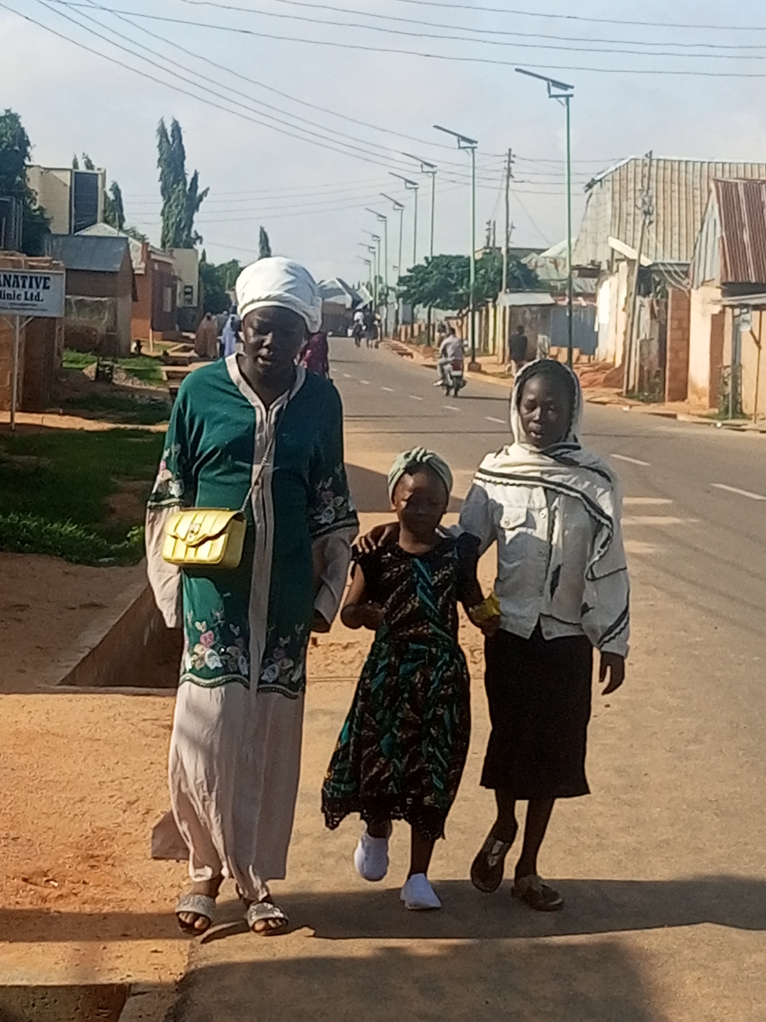 In Tumfure, Nigeria, three individuals walk along a paved road on a sunny day. On the left, an adult, possibly female, wears a long green robe with floral embroidery, a white headscarf, and a yellow purse slung across their chest. They have sandals on their feet. Next to them, a child, possibly female, in a dark patterned dress and a light green head covering, walks with their arm linked with the adult. To the right, another individual, possibly female, wears a light-colored jacket with striped trim, a white and black striped scarf, and a black skirt. They are wearing sandals. The road is lined with buildings, some with corrugated metal roofs, and utility poles with solar panels are visible. The ground is dusty and a shadow is cast by one of the individuals.