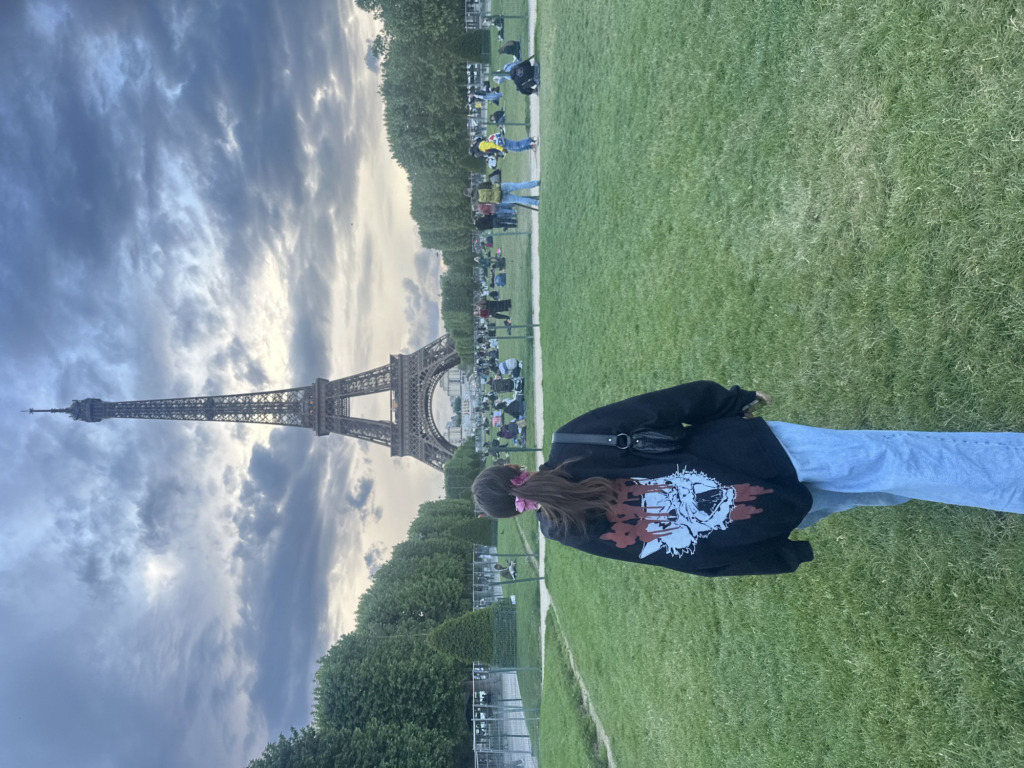A full shot of the Eiffel Tower in Paris, France, and a woman standing on the lawn, facing away from the viewer. The woman is wearing blue jeans, a black sweatshirt with white and red graphics, and a black shoulder bag. Her hair is brown and pulled back with pink hair accessories. The Eiffel Tower is in the background, under a cloudy sky. People are sitting on the lawn in front of the tower. A small fence runs parallel to the path between the woman and the tower. Trees line the path.
