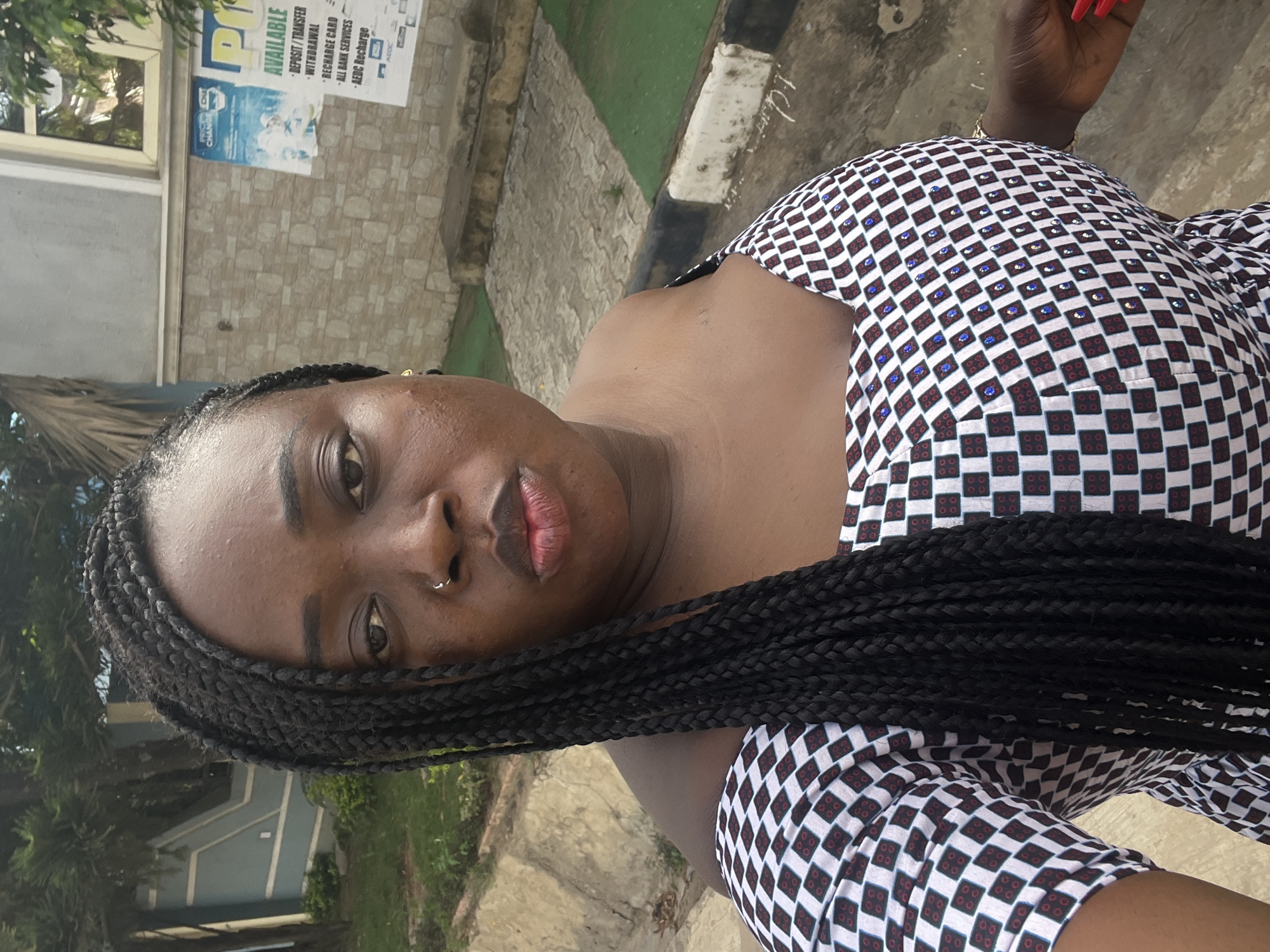 A medium shot depicts a Black woman with dark braids, wearing a black and white checkered patterned top, standing outside. She has a nose ring and her fingernails are painted red.
In the background, there is a building with a poster advertising banking and utility services ("Deposit/Transfer, Withdrawal, Recharge Card, All Bank Services, AEDC Recharge"). The presence of AEDC Recharge suggests the location might be in the city of Abuja, Nigeria, as AEDC stands for Abuja Electricity Distribution Company. There are some trees and greenery visible around the building.