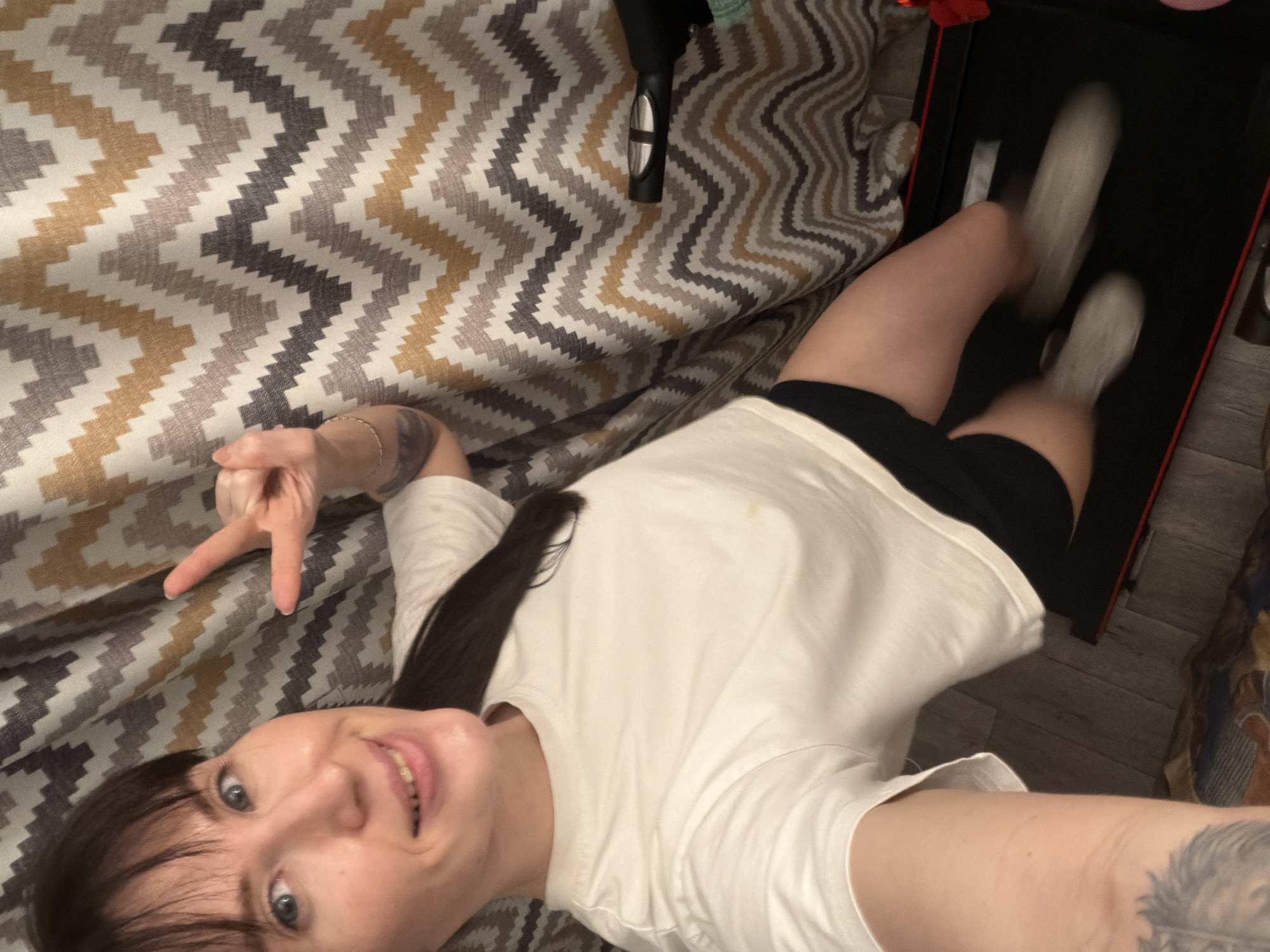A woman is taking a selfie in what appears to be her home. She is wearing a white t-shirt and black shorts. She is giving a peace sign with her left hand. Her feet are on a black treadmill. The background includes a zig-zag patterned blanket and wood flooring. The location is Perm, Russia, based on metadata not present in the visual data.