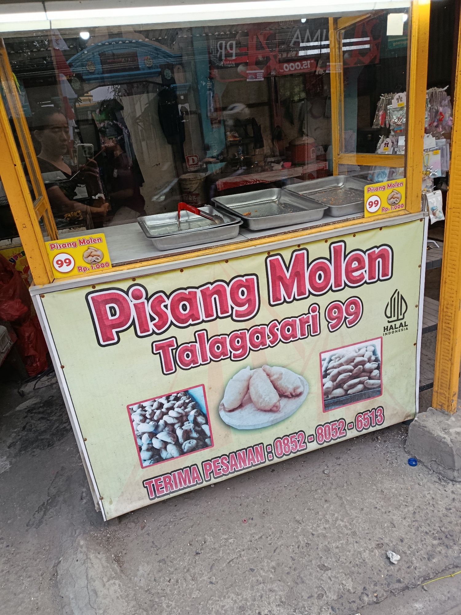 The image shows a street food stall in Balaraja, Indonesia, selling "Pisang Molen" (fried banana fritters). The stall is yellow and has a large banner with the name of the product and images of the fritters. There are also small price tags displayed. In the background, through the glass of the stall, a person is visible holding a phone, possibly taking a picture or recording. The stall is set up on a concrete sidewalk next to a road.