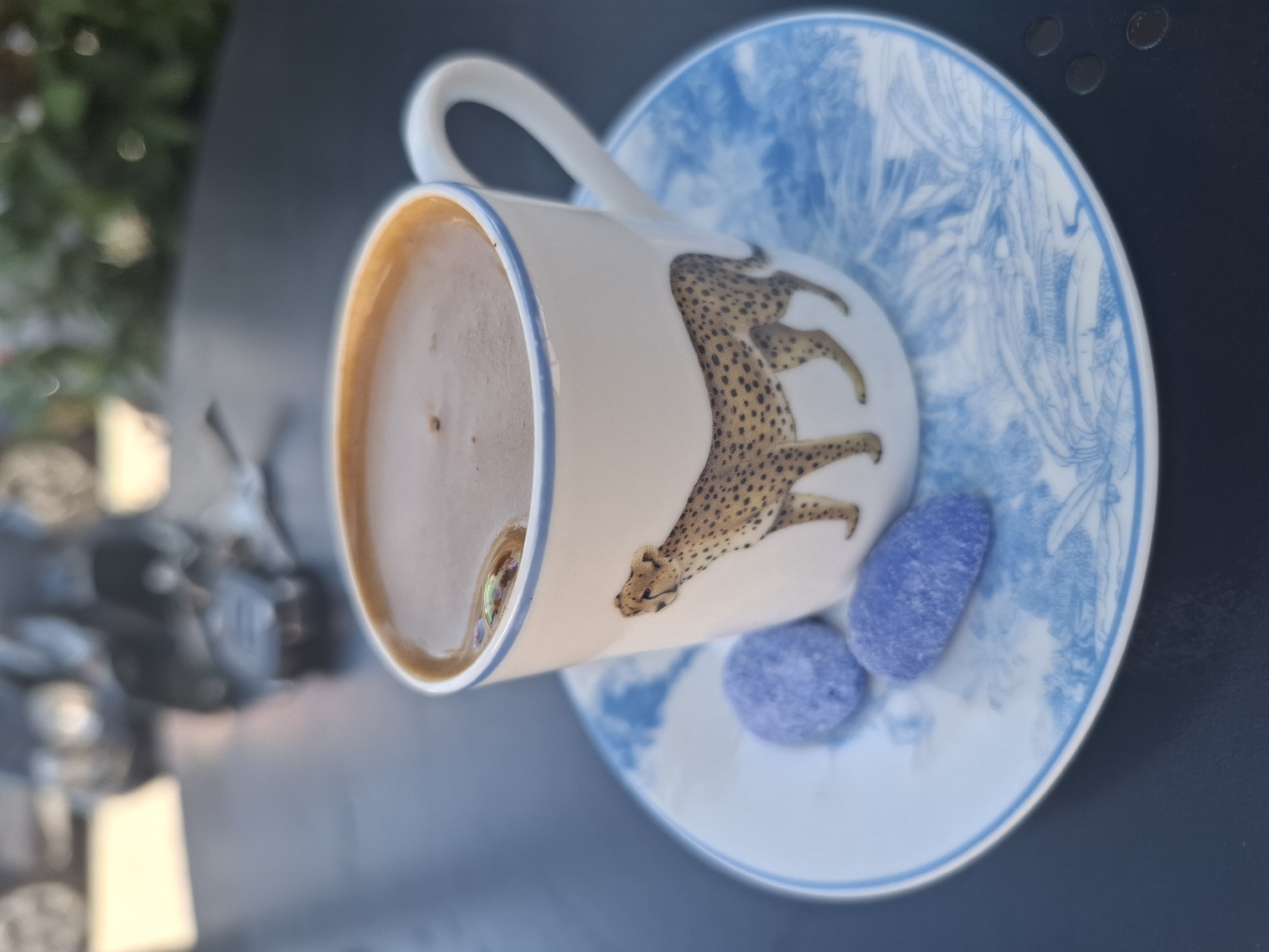 The image shows a close-up of a cup of coffee on a saucer, adorned with a cheetah print and blue floral patterns. Two small, blue candies are placed on the saucer next to the cup. The background is blurred, showing hints of vehicles and greenery, suggesting an outdoor setting. The coffee appears to be dark and frothy, with a few small bubbles on the surface. The cup has a white base with a blue rim, and a handle. The saucer is white with blue decorative elements. The overall impression is a pleasant moment of relaxation, possibly at an outdoor cafe in Fethiye, Turkey, where such decorative cups and traditional sweets might be enjoyed.
