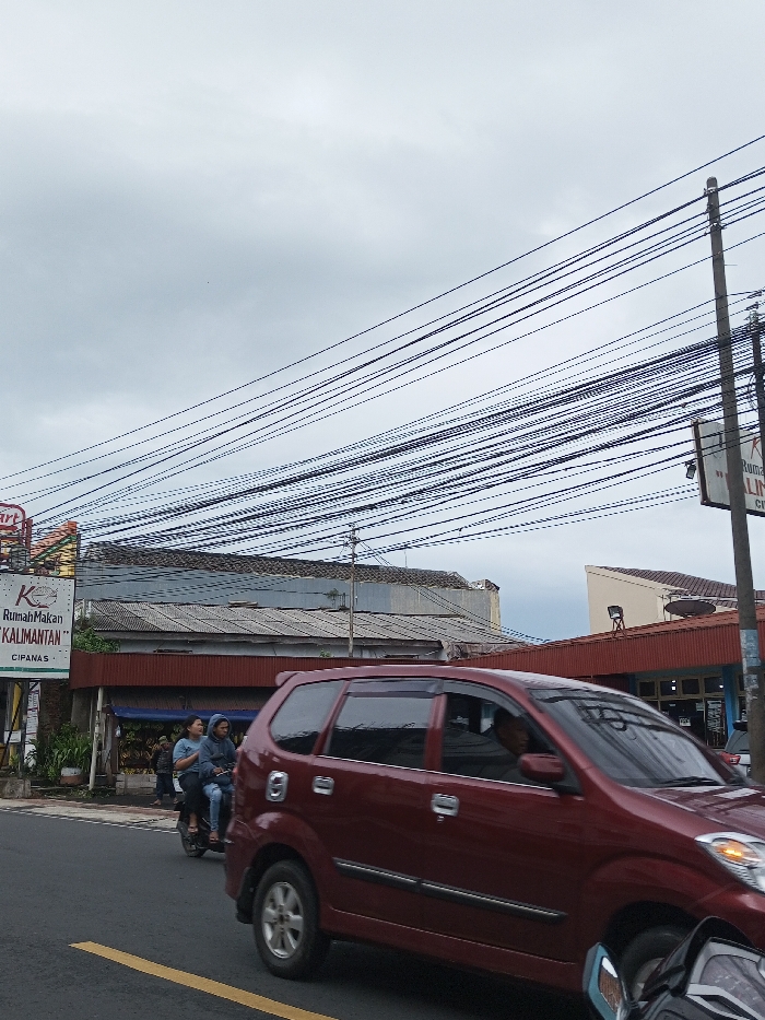 The image depicts a street scene in the city of Pacet, Indonesia, under an overcast sky. The road in the foreground is asphalt, with a faded yellow dividing line partially visible on the left.
A dark red multi-purpose vehicle (MPV) occupies the mid-ground, moving from right to left. A male driver is discernible through the front driver's side window. Directly behind and slightly to the left of the MPV, a dark-colored scooter is in motion, carrying two individuals. The driver wears a dark jacket, and the passenger wears a light blue or grey top. A portion of another motorcycle, black with chrome elements, is visible in the extreme bottom right corner, positioned behind the MPV.
On the left side of the street, a vertical sign for "Rumah Makan Kalimantan" (Kalimantan Restaurant) with "CIPANAS" printed below is visible. Adjacent to this sign, greenery lines the roadside, preceding buildings. The primary building on the left features a corrugated metal roof and a reddish-brown facade below, likely a commercial establishment. Further to the right, another building with a similar reddish-brown facade and a contrasting light-colored roof is visible.
A dense array of black overhead electrical or utility wires crisscrosses the upper portion of the frame, originating from the top left and extending diagonally towards the mid-right. A concrete utility pole stands on the right side, supporting these wires and a partially obscured sign. In the background on the right, a building with a sloping light-colored wall and a brown roof is present. The overall lighting is diffuse, characteristic of an overcast day.