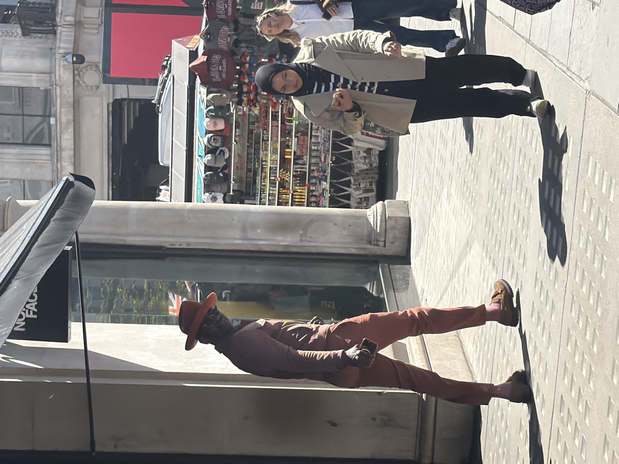 This full-length, vertically oriented image, captured under bright daylight, depicts a bustling street scene in what appears to be a commercial or tourist area of London, United Kingdom.
In the foreground, to the left of the frame, a man with dark skin stands on a patterned grey sidewalk. He is dressed in a striking monochromatic burnt orange or reddish-brown suit, consisting of a jacket, trousers, and a fedora-style hat. He wears brown shoes, revealing vibrant pink socks, and is holding a black smartphone in his right hand. His face is largely shadowed by the brim of his hat.
Further back and to the right, another individual, a woman, is visible. She wears a black hijab, a horizontal black and white striped top, black trousers, and a light beige trench coat. Her expression appears neutral to slightly pleasant as she looks towards the viewer. Behind her, another blonde-haired woman in a white shirt and jeans is partially visible.
The background features several street vendor stalls laden with merchandise. Visible items include various colored caps and red hooded sweatshirts, some clearly displaying the word "LONDON," indicating a tourist destination. Architectural details of a stone building with windows are seen behind the man. Near the foreground on the left, a partially visible black sign or stand features the white text "NO FACE," suggesting it is part of a "THE NORTH FACE" logo.
The scene is brightly lit by strong sunlight, casting long, distinct shadows of the people and an overhead awning onto the patterned sidewalk, suggesting it is daytime. The combination of "LONDON" merchandise, the visible Union Jack flag (faintly reflected in a window), and typical city architecture strongly confirms the specified location context of London, United Kingdom.