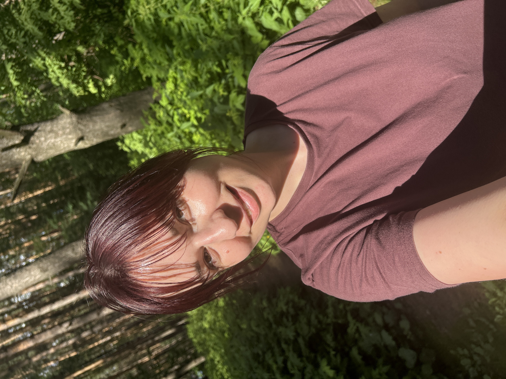 A young woman with dark, auburn hair and bangs smiles at the camera in a selfie taken outdoors. She is wearing a dark magenta t-shirt. The background consists of a dense forest with tall trees and lush green foliage, bathed in dappled sunlight. The scene suggests a recreational area, possibly a park or trail within the city of Chaikovsky, Russia, given the prevalence of similar forested landscapes in the region. The lighting indicates it is daytime, likely late afternoon due to the long shadows cast by the trees. There are no other individuals or notable objects in the frame.