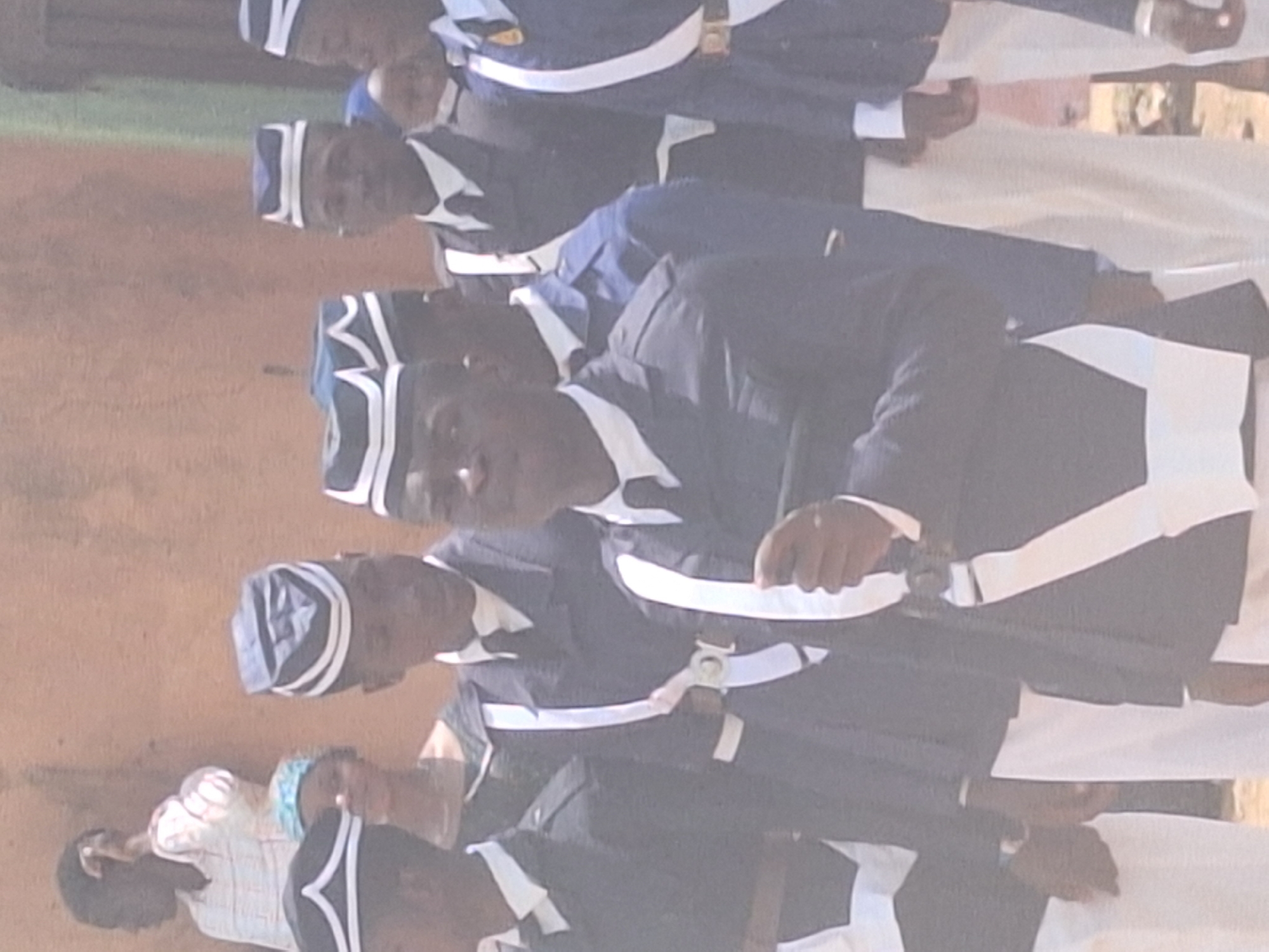 A group of men are standing in a row, wearing dark-colored uniforms with white trim and hats with a white design. They are in Sabon Tasha, Nigeria, holding what appear to be ceremonial swords. One person is visible to the left of the group and appears to be wearing a light-colored shirt. The background appears to be a dusty, outdoor area.