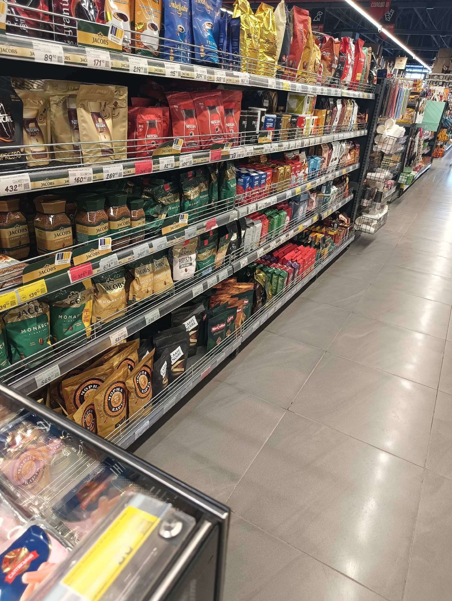 This image presents a forensic view of a supermarket aisle in Maiaky, Ukraine, dedicated to coffee products. Multiple metal shelving units display a wide array of coffee brands, including Jacobs, Nescafe, Monarch, Ambassador, Ristretto, and Carte Noire, packaged in various formats such as bags, pouches, and glass jars. Price tags, denominated in Ukrainian Hryvnia, are affixed to the shelves below each product.
The top shelves feature large bags of ground and whole bean coffee alongside glass jars of instant coffee, including Jacobs Cronat Gold. The middle shelves are stocked with Nescafe Gold instant coffee in pouches, distinctive red Nescafe packages, and other boxed and bagged coffee varieties. The lower shelves predominantly display glass jars of Jacobs Cronat Gold and Monarch instant coffee, large bags of Jacobs Monarch, and black and gold bags of coffee labeled "ЧОРНА КАРТА" (Black Card).
The floor is composed of light grey rectangular tiles, exhibiting reflections from overhead fluorescent strip lighting, which is also visible on the ceiling. To the right of the primary coffee display, additional store shelving is partially visible, containing items such as plastic straws and disposable cups arranged in wire baskets. In the bottom-left foreground, a segment of a refrigerated display case is observable, containing packaged food items, with a blue package marked "САЛТІВСЬКИЙ" partially legible. No individuals are distinctly visible within the immediate area of focus.