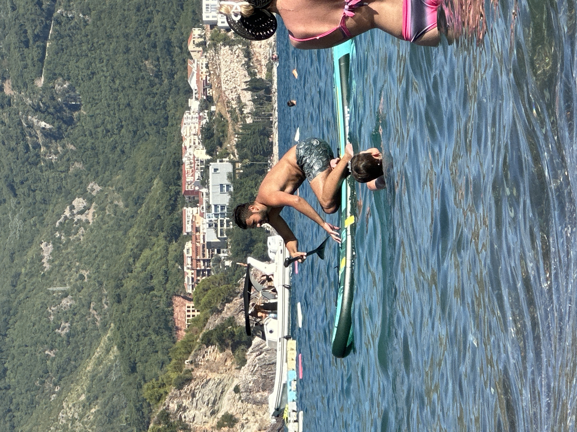 The image shows a group of people in the water near a coastline. A man, shirtless and wearing patterned swim trunks, is on a paddleboard, leaning forward as if to grab something or adjust his position. Another person, a child, is also on the paddleboard, with their head visible. A portion of another person's lower body, wearing a pink and gray bikini, is visible at the top of the frame, suggesting they are also in the water or on a nearby vessel.

The background features a steep, tree-covered hillside with buildings clustered along its lower slopes, indicative of a coastal town. A white boat is docked or anchored near the shore. The water is a clear blue, reflecting the sunlight, and the overall scene appears to be set on a sunny day, likely during daylight hours. The mood seems relaxed and recreational, consistent with activities like paddleboarding. The location context of Boreti, Montenegro, aligns with the coastal scenery depicted. No visible text is present in the image.