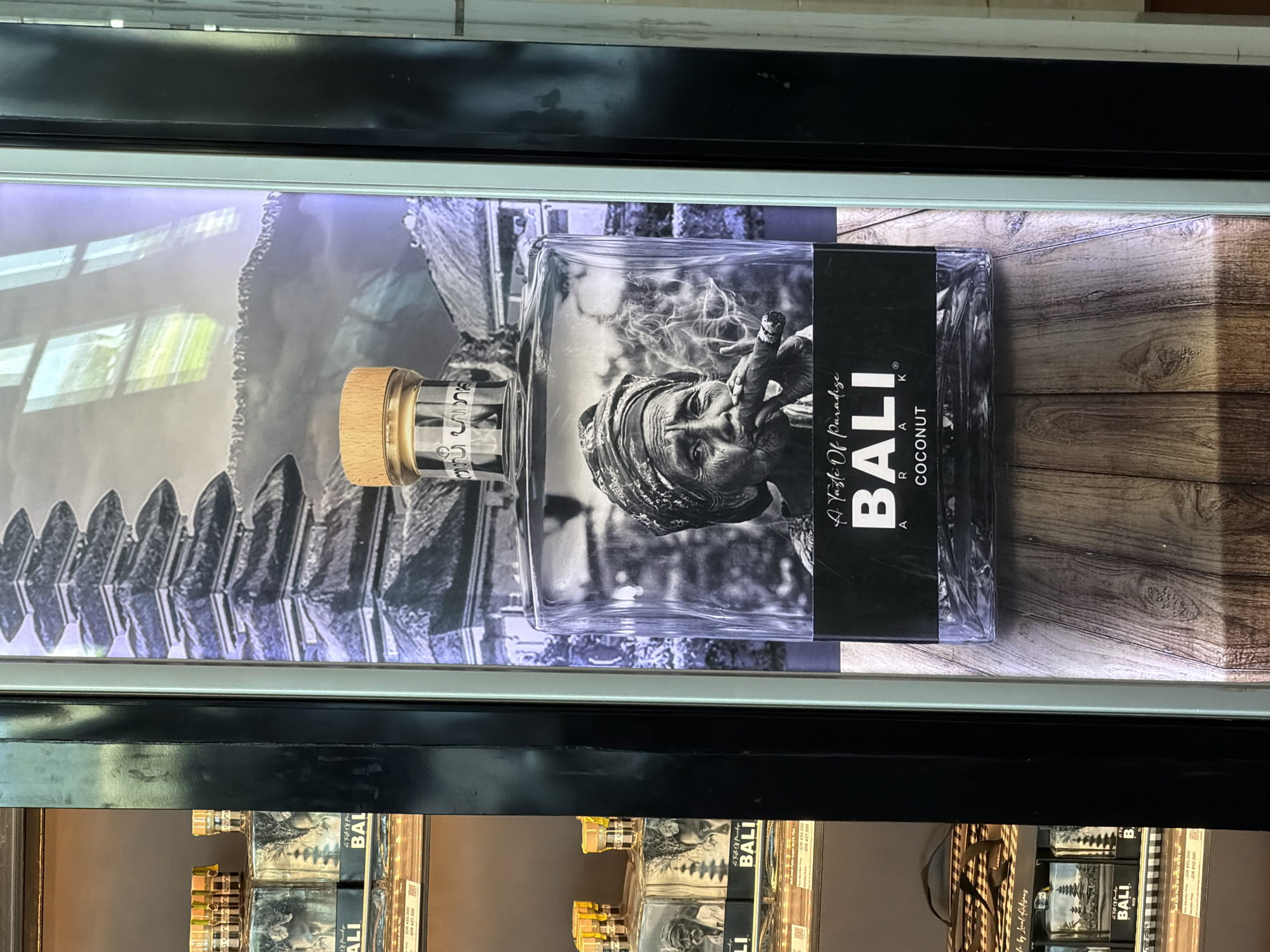 The image shows a shelf with multiple bottles of "Bali Coconut" perfume. Above the bottles is a display advertisement for the same perfume, featuring a bottle against a backdrop of a Balinese temple and a black-and-white portrait of an elderly woman smoking a cigar. The image was taken in Kuta, Indonesia.