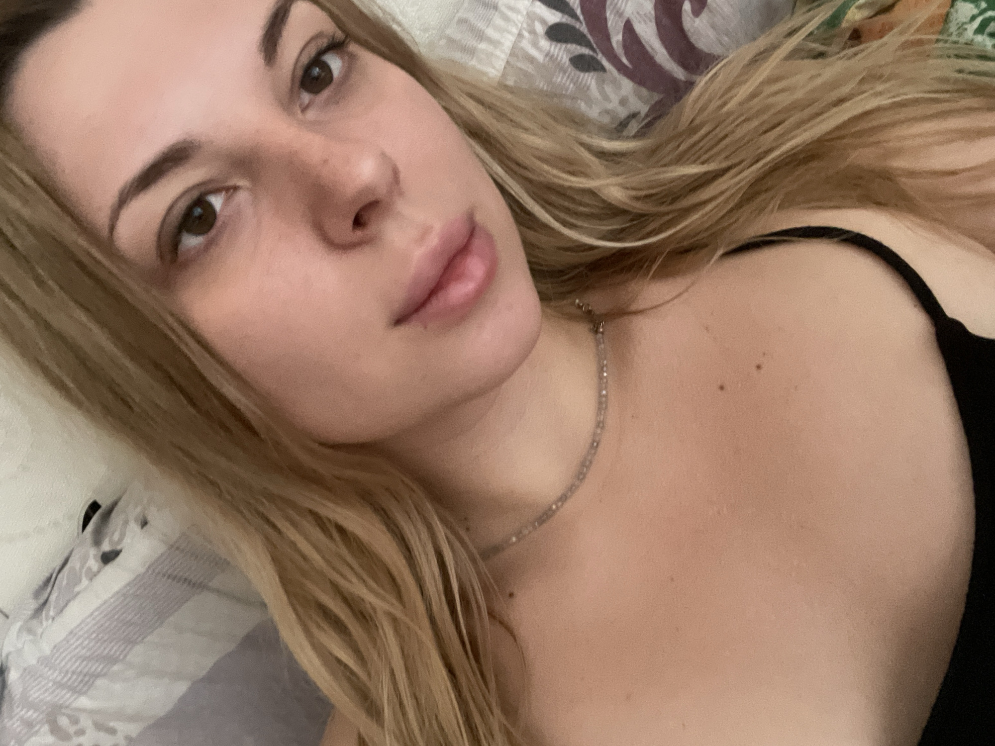 A close-up, head and shoulder selfie of a blonde woman with fair skin, brown eyes, and light brown eyebrows is shown. She is wearing a black bra and a delicate necklace. Her hair is long and wavy, falling around her face and shoulders. The background consists of a striped pillow and bedding. The image quality is moderate, with some pixelation visible. There is no discernible city or specific location information, including Shcherbinka, Russia.