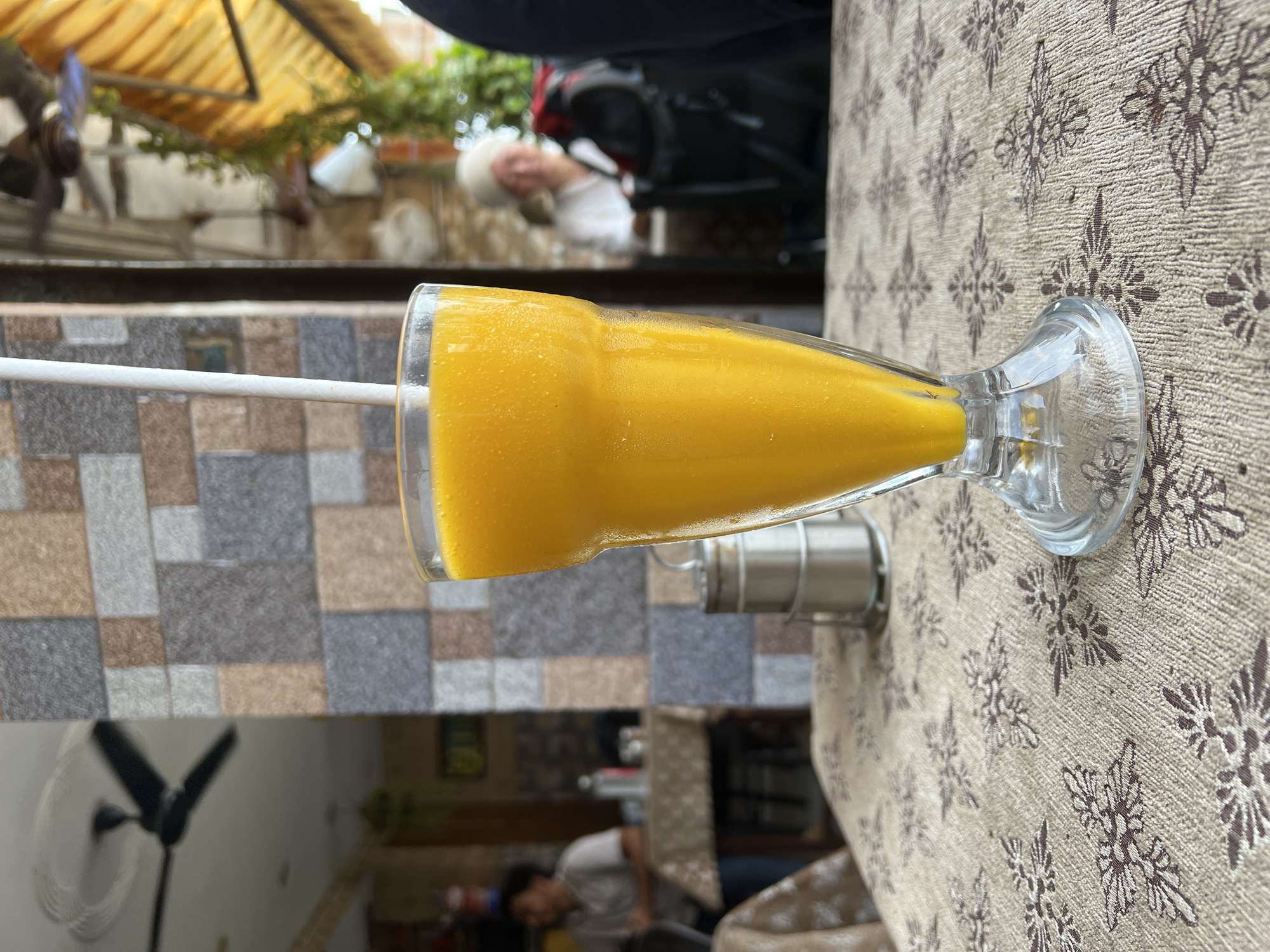 A tall glass of bright orange mango lassi sits on a patterned tablecloth. A white straw extends from the drink. In the background, a wall with colorful tiles is visible, along with a ceiling fan. Further back, a person is seated at a table, and another person is partially visible in the far background. The overall setting suggests an outdoor cafe or restaurant. This scene is located in Delhi, India.
