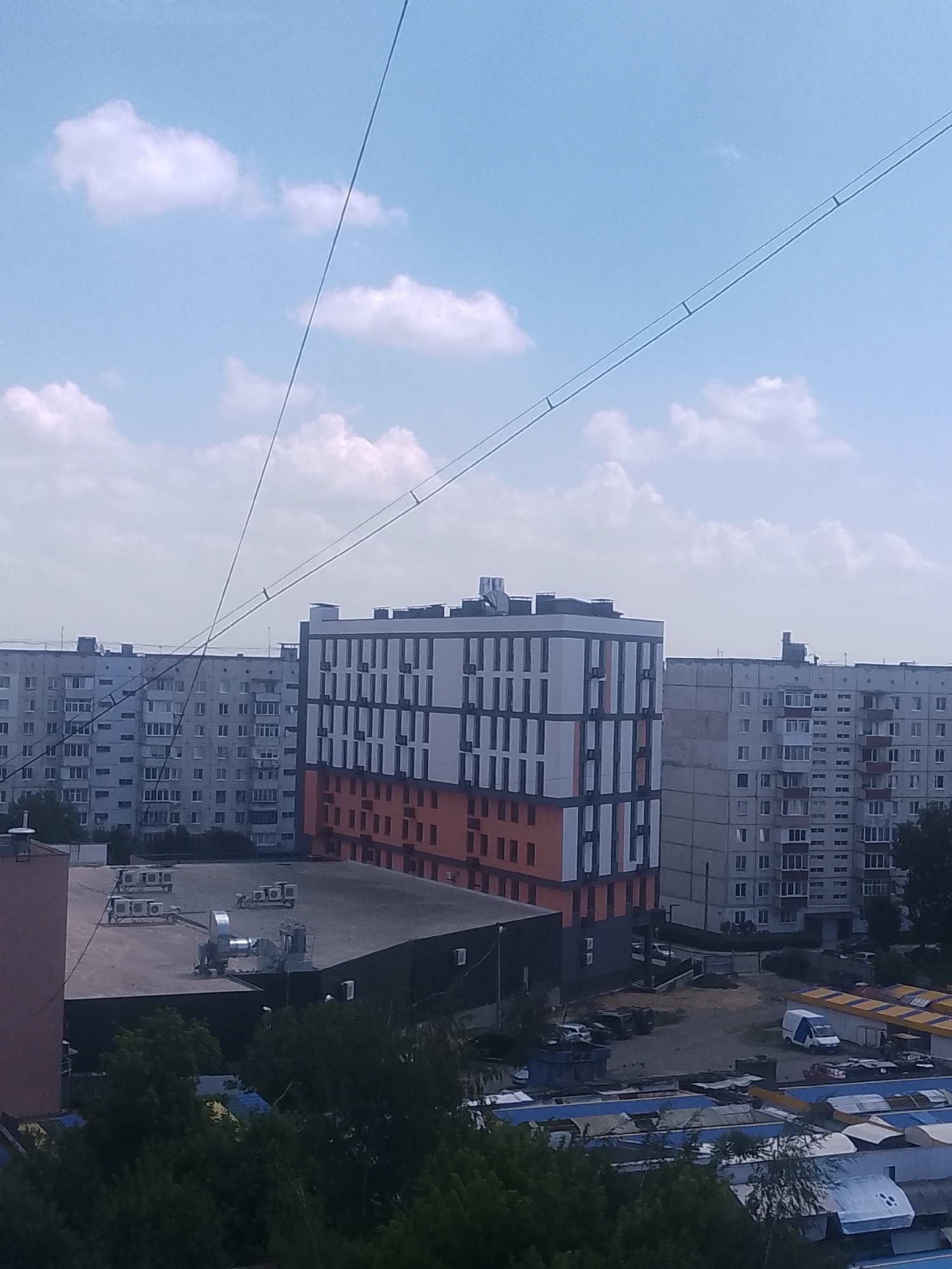This is an aerial photograph taken from a high vantage point in Chernivtsi, Ukraine, looking across a cityscape. The sky is partly cloudy with a pale blue hue. Several buildings are visible, including a modern apartment building with orange and white geometric accents, and a series of older, grey apartment blocks. In the foreground, a flat, dark-colored rooftop area is visible, equipped with multiple air conditioning units. Beyond this, the photograph shows a collection of market stalls or covered outdoor areas with yellow and blue awnings, and further in the distance, more residential buildings. Several vehicles, including a white van, are parked in the area with market stalls. The scene appears to be an urban environment on a clear day.