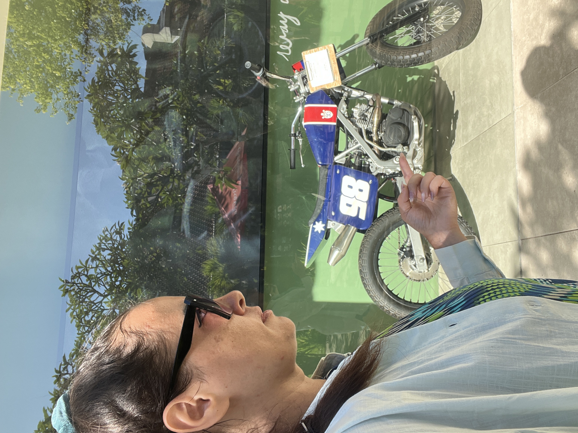 In Kuta Selatan, Indonesia, a woman with dark hair pulled back and wearing sunglasses is standing in front of a display. She is wearing a light blue collared shirt over a patterned top. Her right hand is extended, with her index finger pointing towards a blue and white flat track motorcycle with the number "98" on its side. The motorcycle is positioned in front of a green wall that has faint white text, possibly "eray a". Behind the motorcycle and the woman, trees and a blue sky are visible, suggesting an outdoor or semi-outdoor setting. The ground is paved with light-colored square tiles.