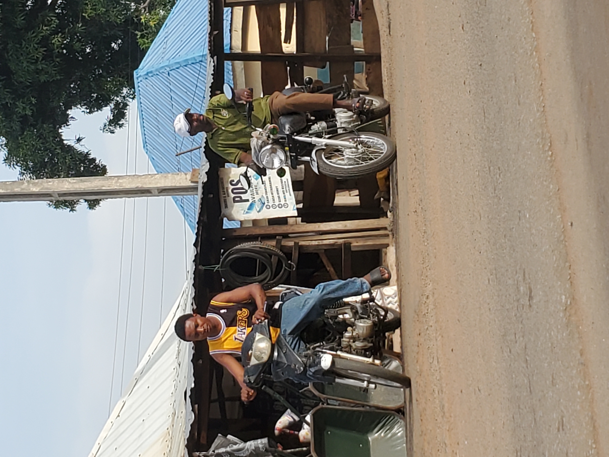 This image depicts a vibrant roadside scene in Mararaba, Nigeria, under bright daylight. Two men are featured, each seated on a motorcycle by an informal, open-air stall or workshop.
On the right, a man wearing a light green shirt, brown trousers, and a white cap is perched on a silver and chrome motorcycle, looking straight ahead. Beside him is a white sign with prominent blue text reading "POS AVAILABLE HERE." Below this, smaller text lists various services: "CASH TRANSFER," "CASH WITHDRAWAL," "SEND MONEY," "RECEIVE MONEY," "AIRTIME RECHARGE," "DATA SUBSCRIPTION," "BETTING WALLET FUNDING," and "BILL PAYMENTS." The structure behind him has a blue corrugated metal roof and wooden supports.
On the left, another man is seated on a darker motorcycle. He is wearing a yellow and black "LAKERS" basketball jersey, blue jeans, and sandals, and is looking towards the camera with a neutral expression. Behind him, another section of the stall features a silver corrugated metal roof.
The ground on the right appears to be a dirt or unpaved road. In the background, there's a large green tree against a clear, light blue sky, and a concrete utility pole is visible. Around the makeshift structures, items such as tires and a green wheelbarrow-like object are present. The scene suggests a typical bustling day at a local roadside business.